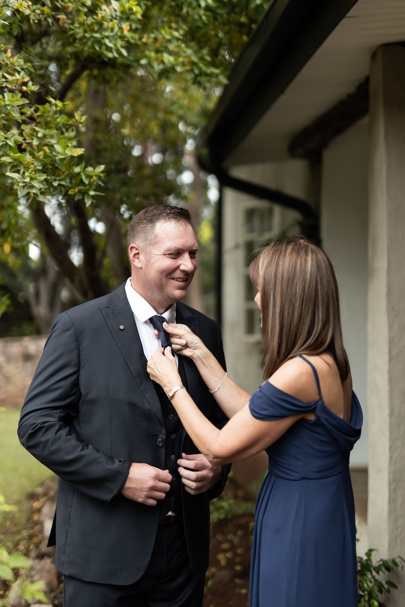 A woman in a navy dress adjusts a boutonniere on a man's black suit jacket outdoors.