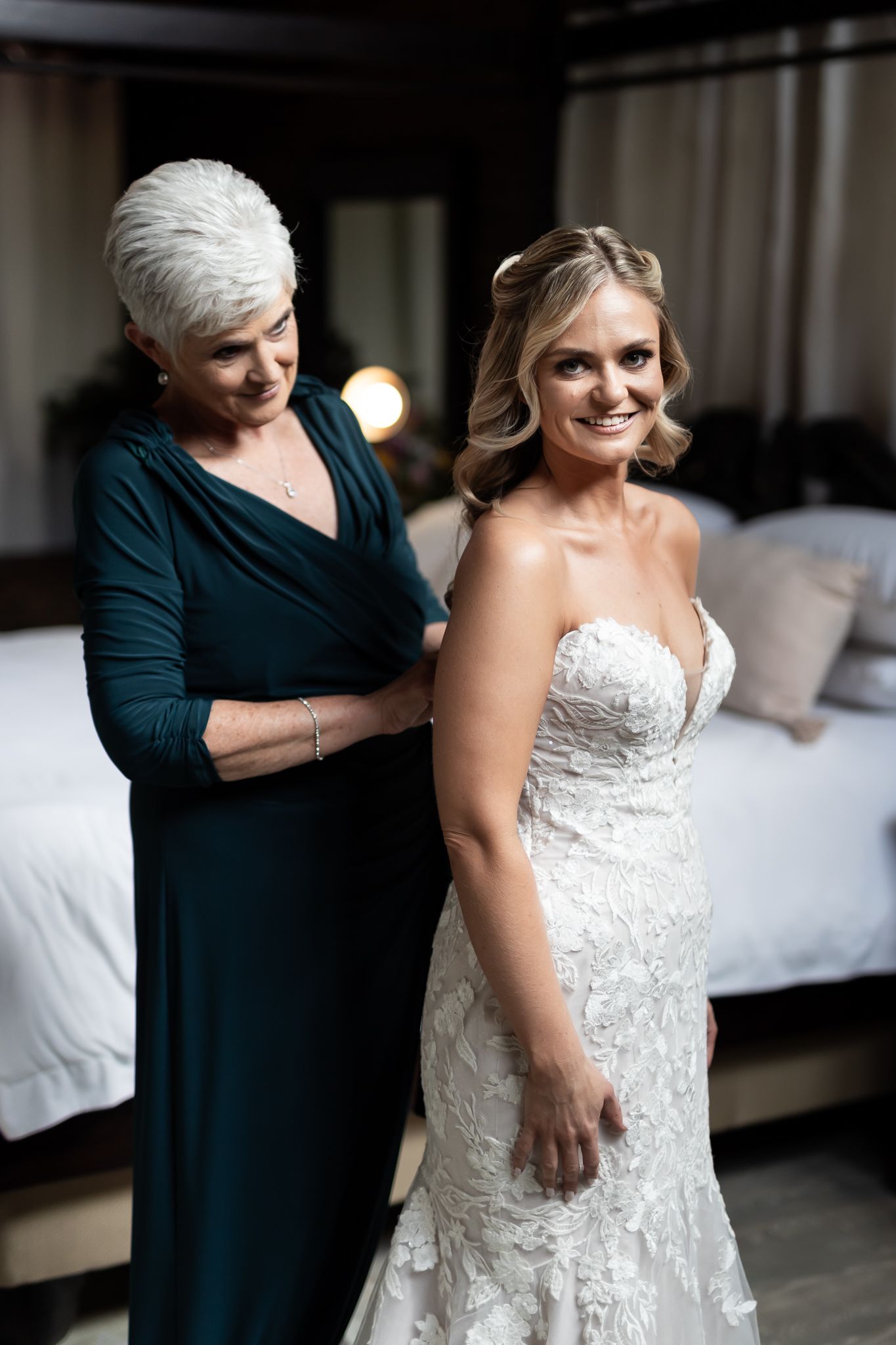 A woman in a teal dress helps a bride get ready in an elegant lace wedding gown in a bedroom setting.