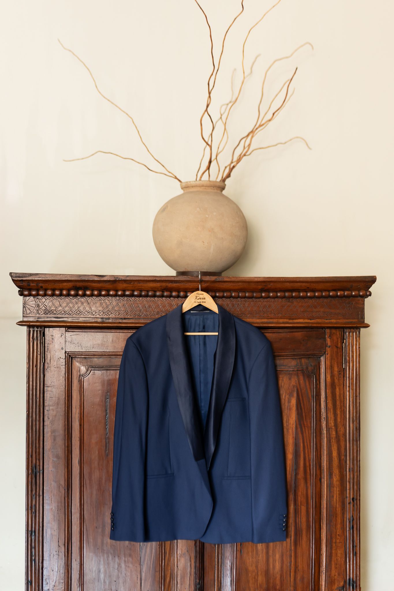 Navy blue suit jacket hanging on wooden wardrobe with decorative vase above.