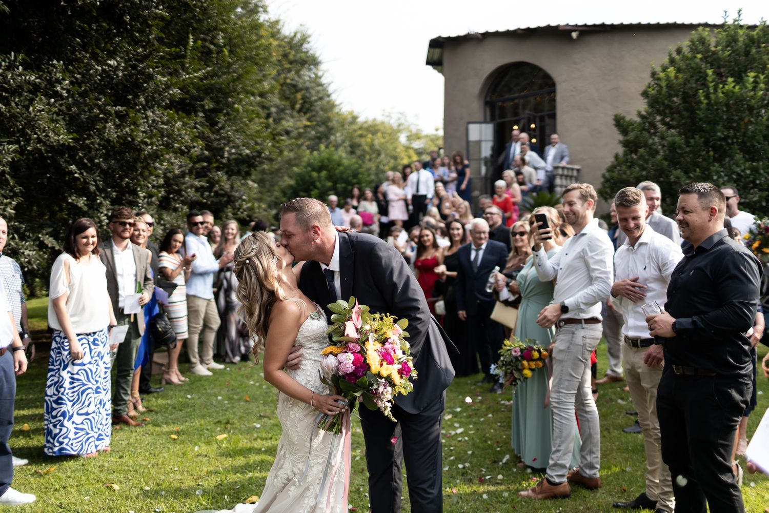Newlyweds with bright floral bouquet greet guests on church lawn.