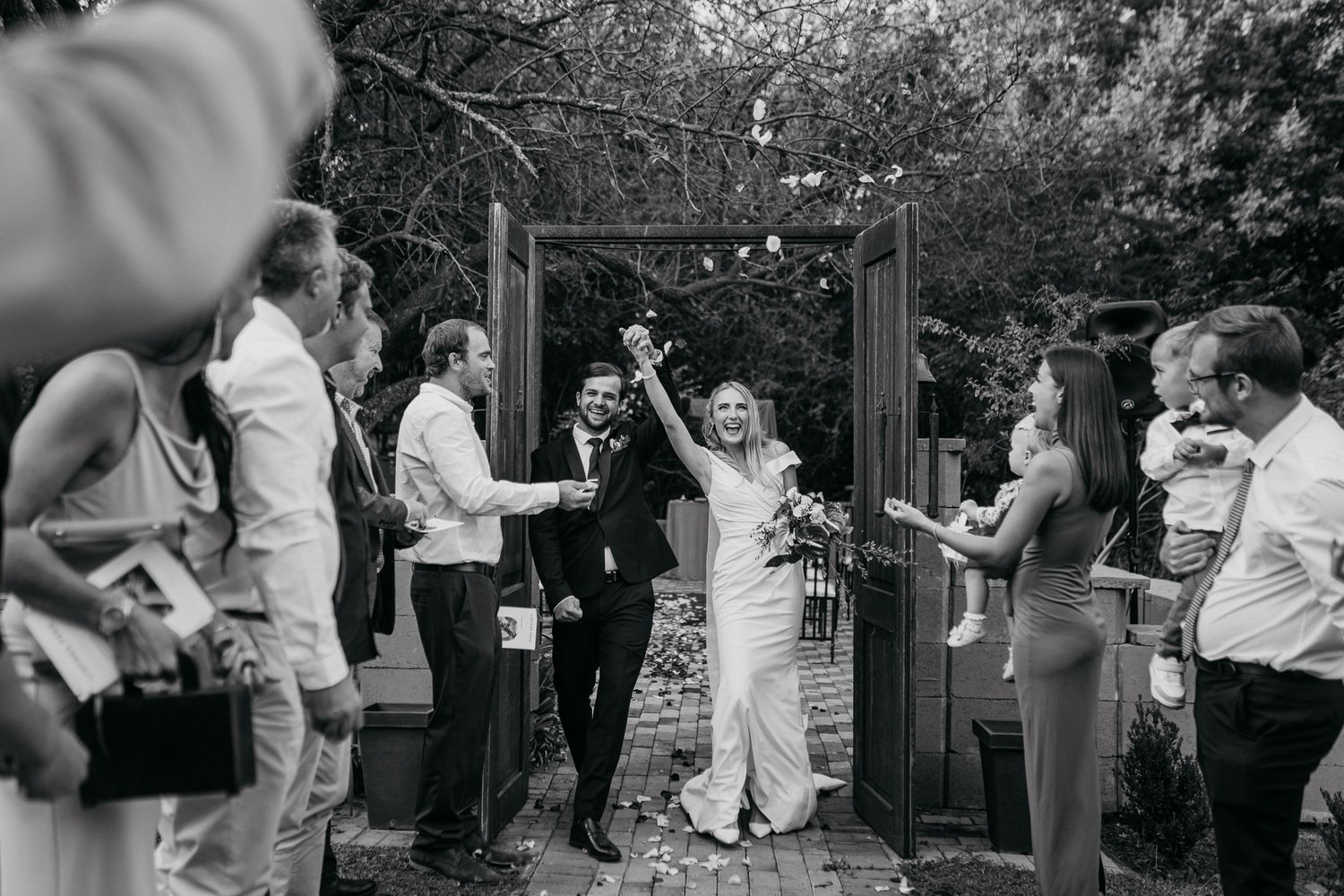 Black and white photo of wedding celebration with guests cheering as couple exits through garden arch.