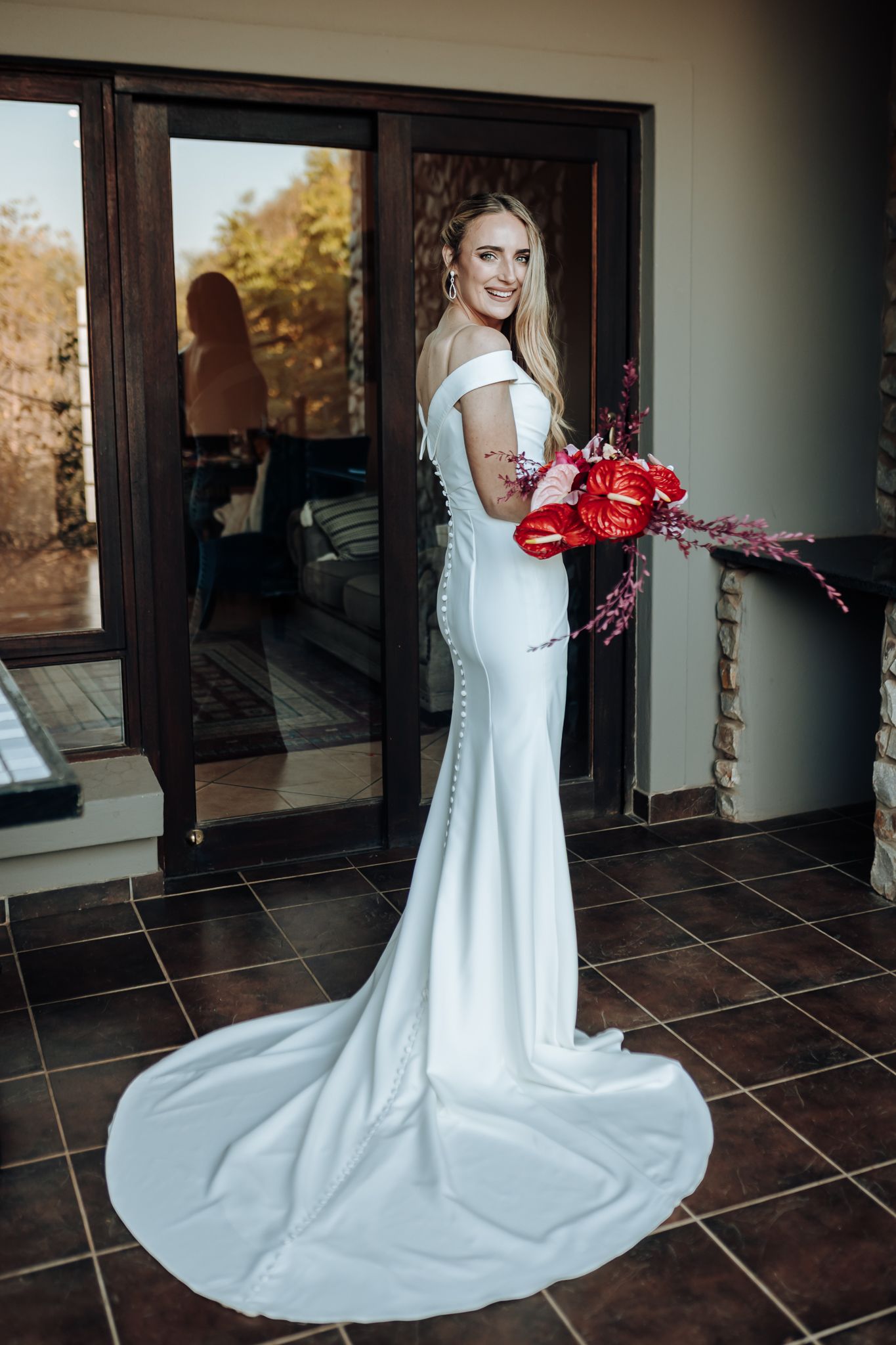 Full-length portrait of bride in fitted dress with cathedral train holding red flowers.