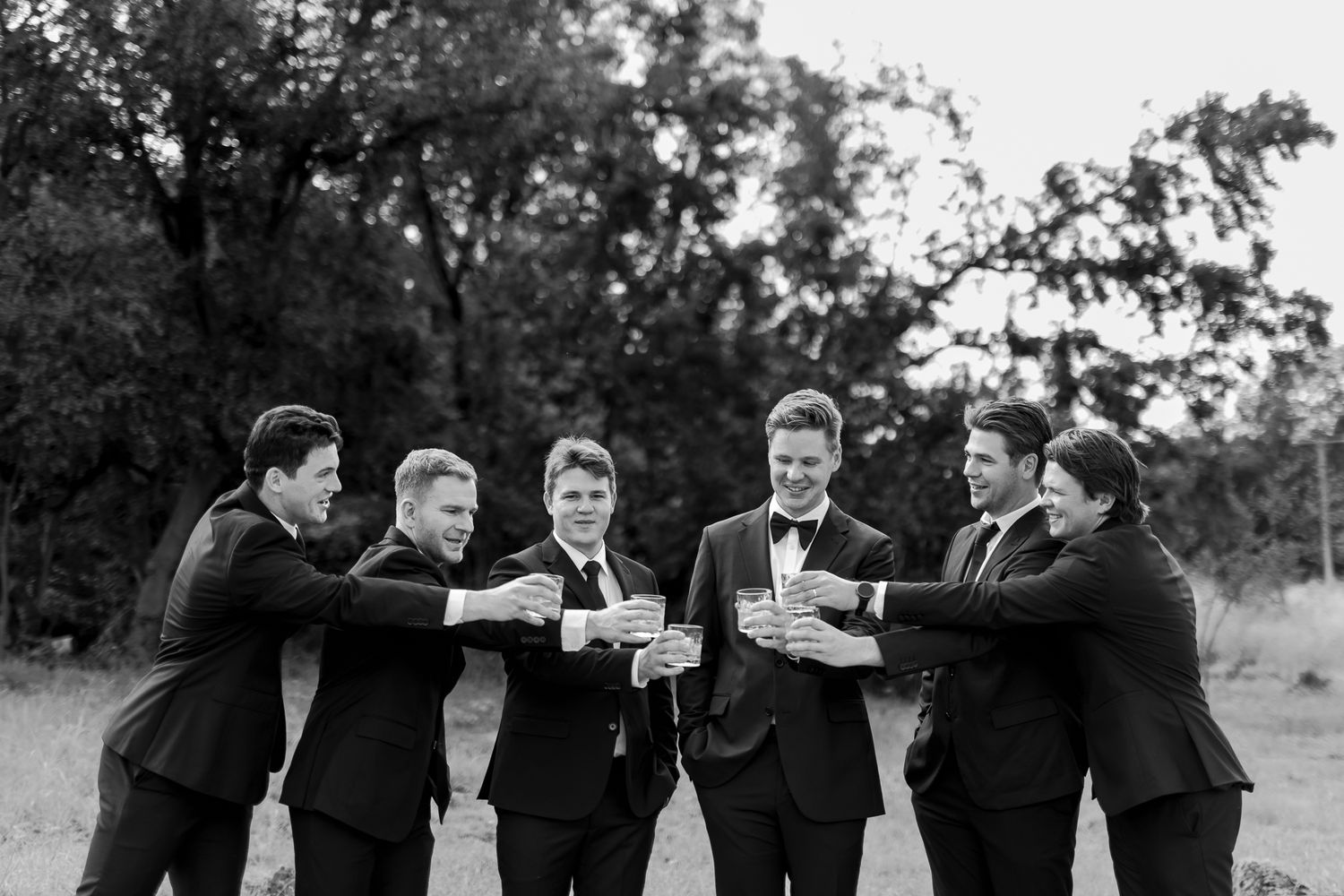 Groomsmen in black suits toasting drinks together outdoors in black and white photography.