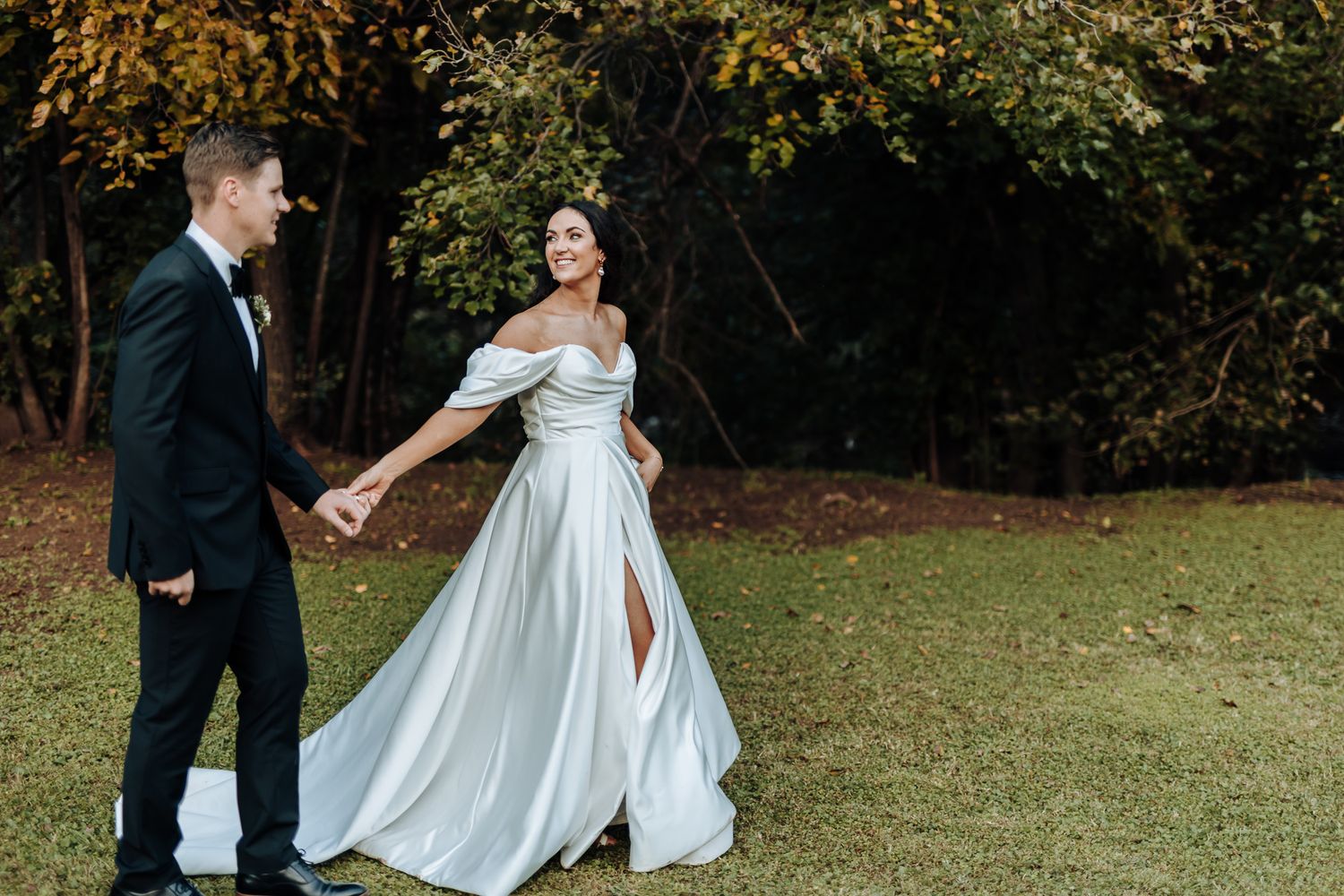 A couple in wedding attire share a playful moment on grassy lawn with autumn trees in background.