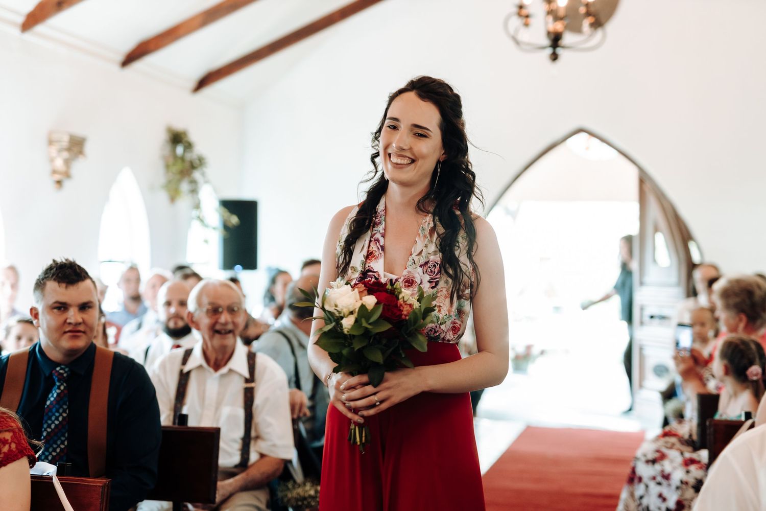 Bridesmaid smiling while holding floral bouquet during chapel ceremony.