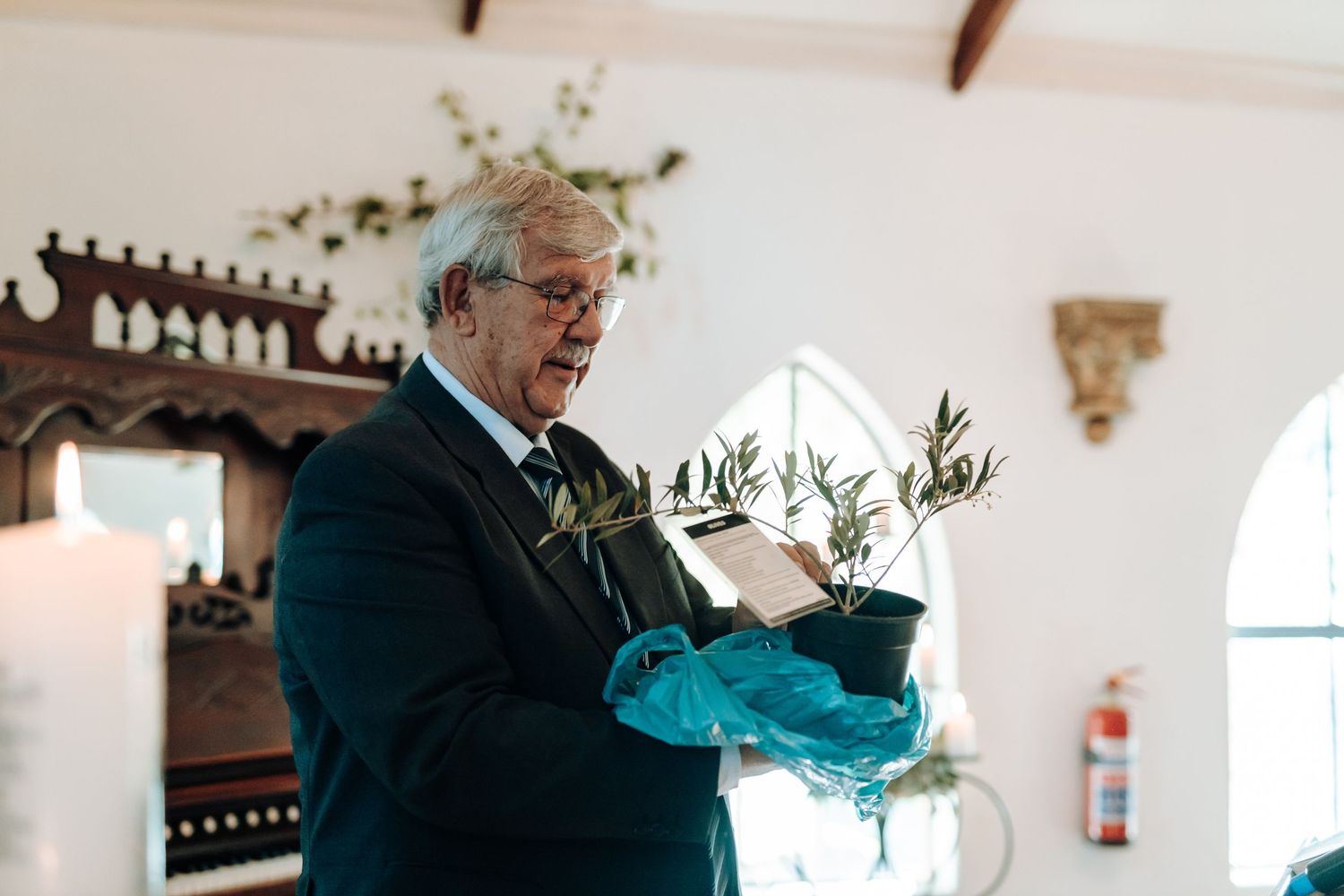 Wedding officiant in navy suit holds decorative arrangement during indoor ceremony.