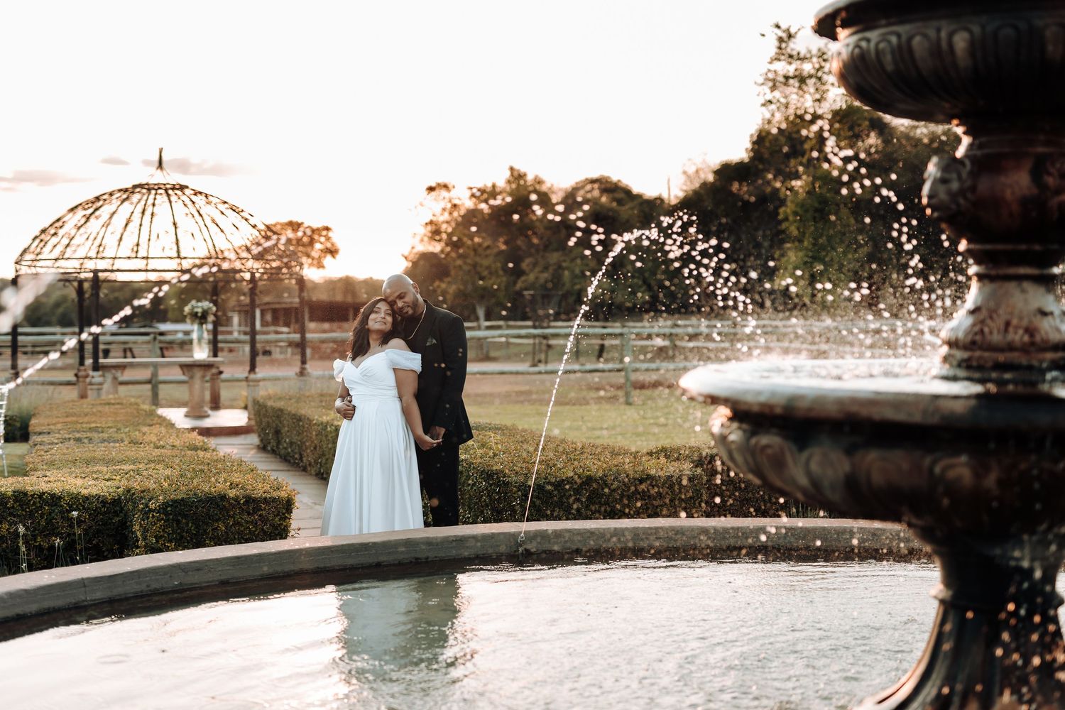 A romantic wedding couple stands by an ornate fountain with water spraying in a garden at sunset.