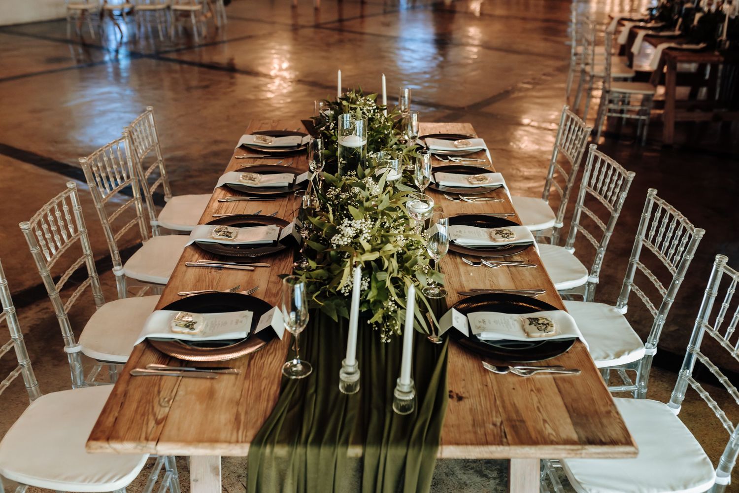 Long dining table set with greenery runner and white place settings for wedding reception.