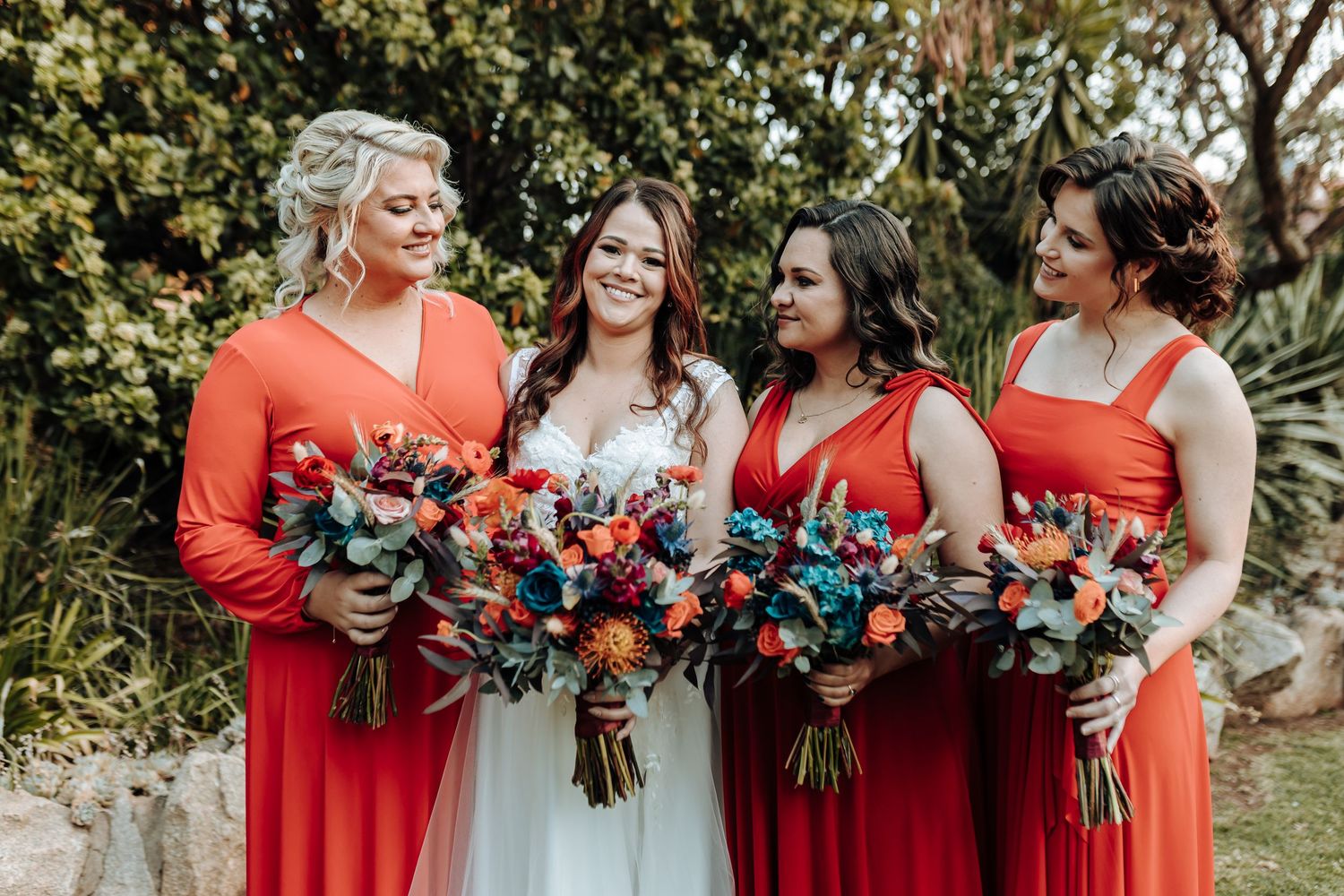 Bridal party poses outdoors in coral and red dresses holding vibrant floral bouquets.
