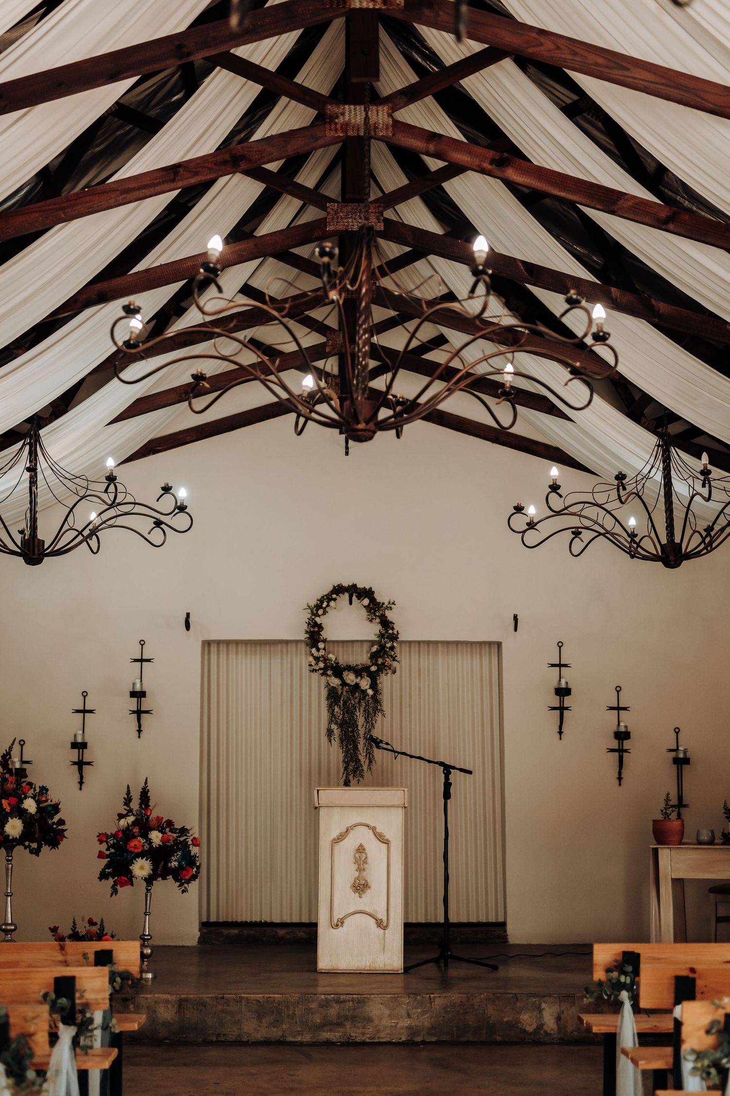 Chapel interior featuring exposed wooden beams, ornate chandeliers, and an altar decorated with a wreath and flowers.