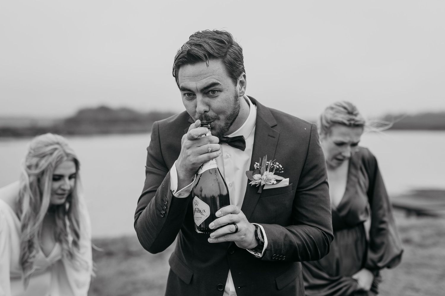 Black and white photo of groom opening champagne bottle outdoors.