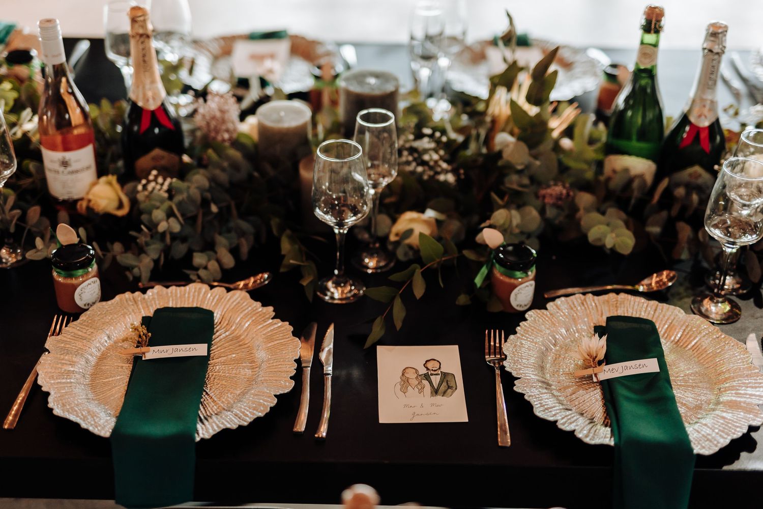 Elegant table setting with green napkins, gold chargers, and wine bottles surrounded by greenery.
