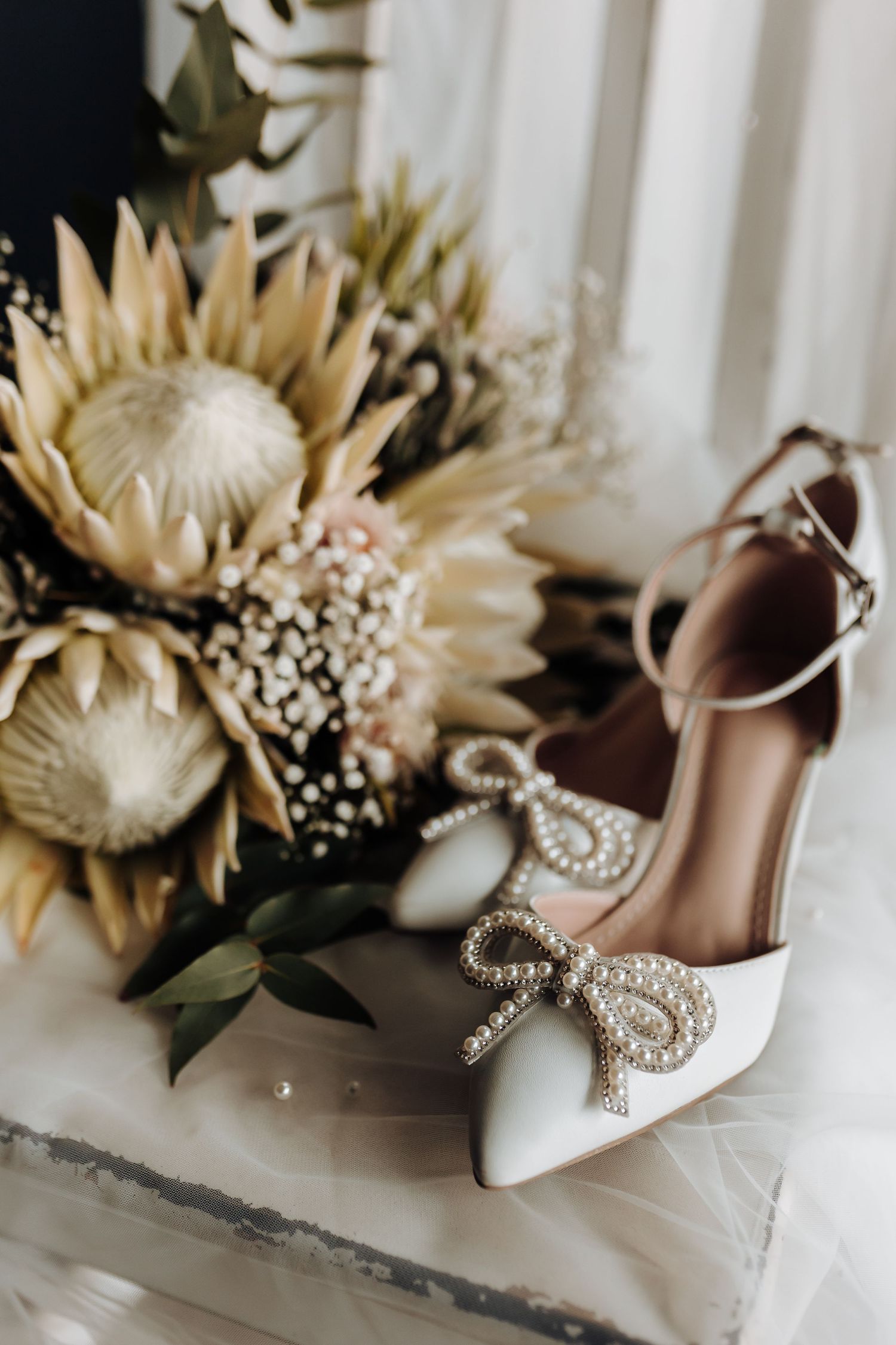 Bridal heels with crystal bow detail displayed next to dried protea flowers and baby's breath.