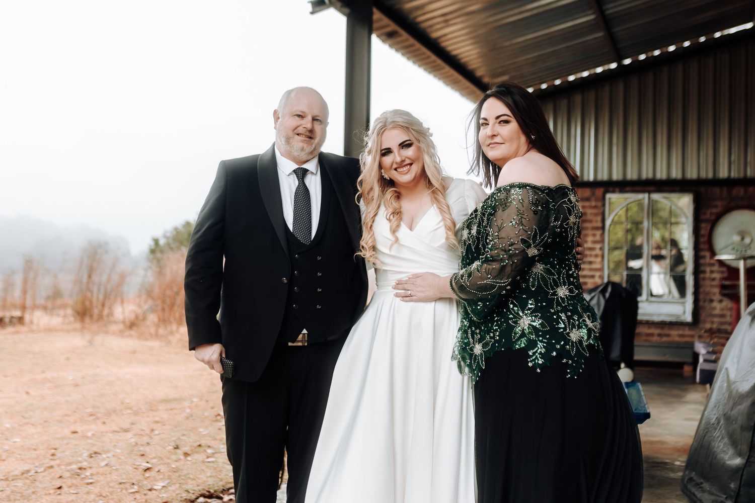 Wedding party portrait featuring the couple with attendant in black dress holding greenery bouquet.