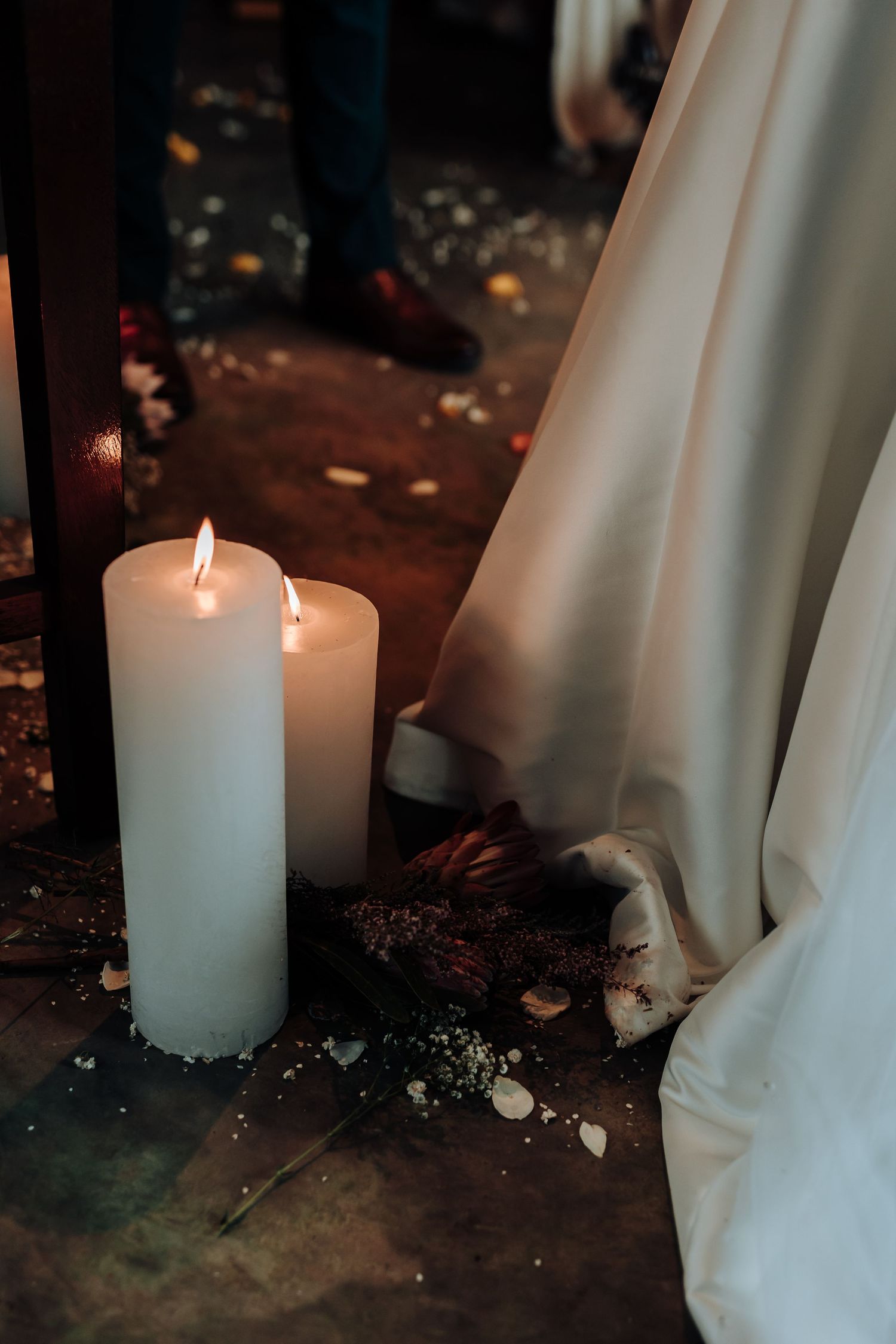 Lit white pillar candles next to flowing wedding dress fabric on rustic floor with scattered petals.