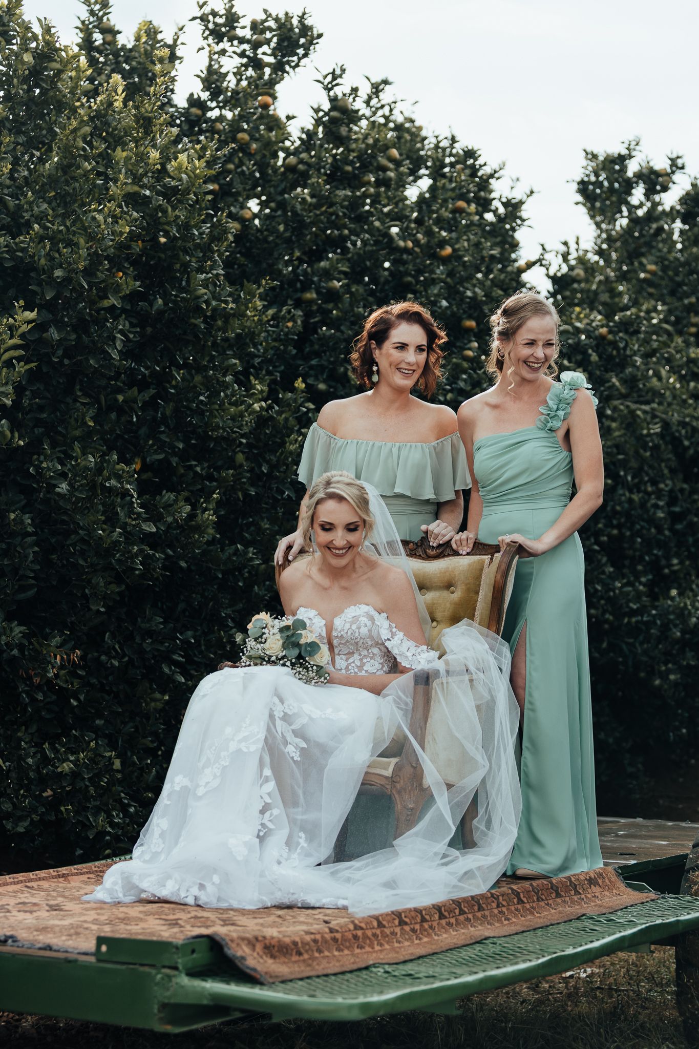 Three attendants in mint green dresses pose on a vintage tractor platform at an outdoor wedding.