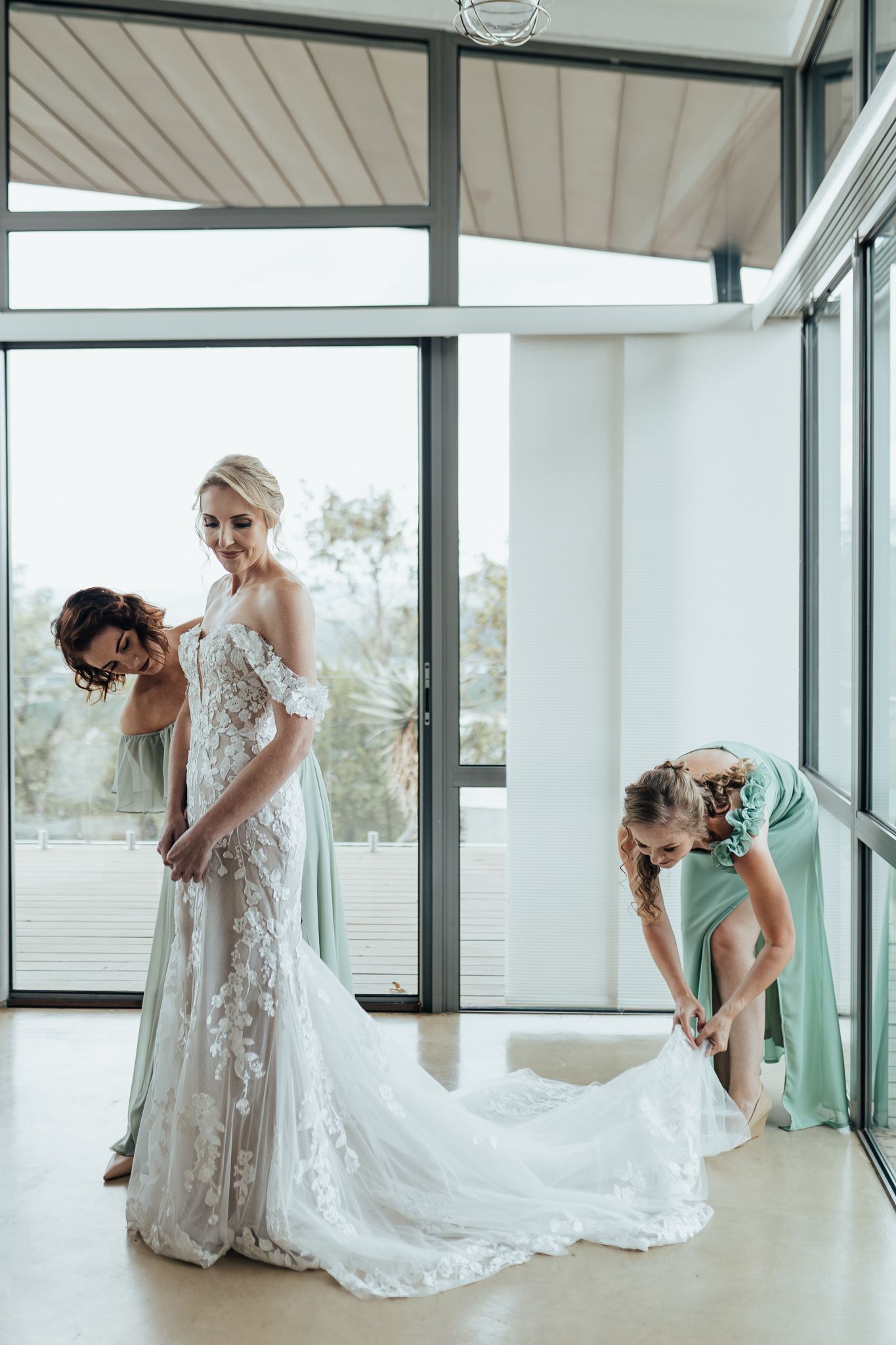 Final adjustments being made to a flowing lace wedding gown train in a modern room with large windows.