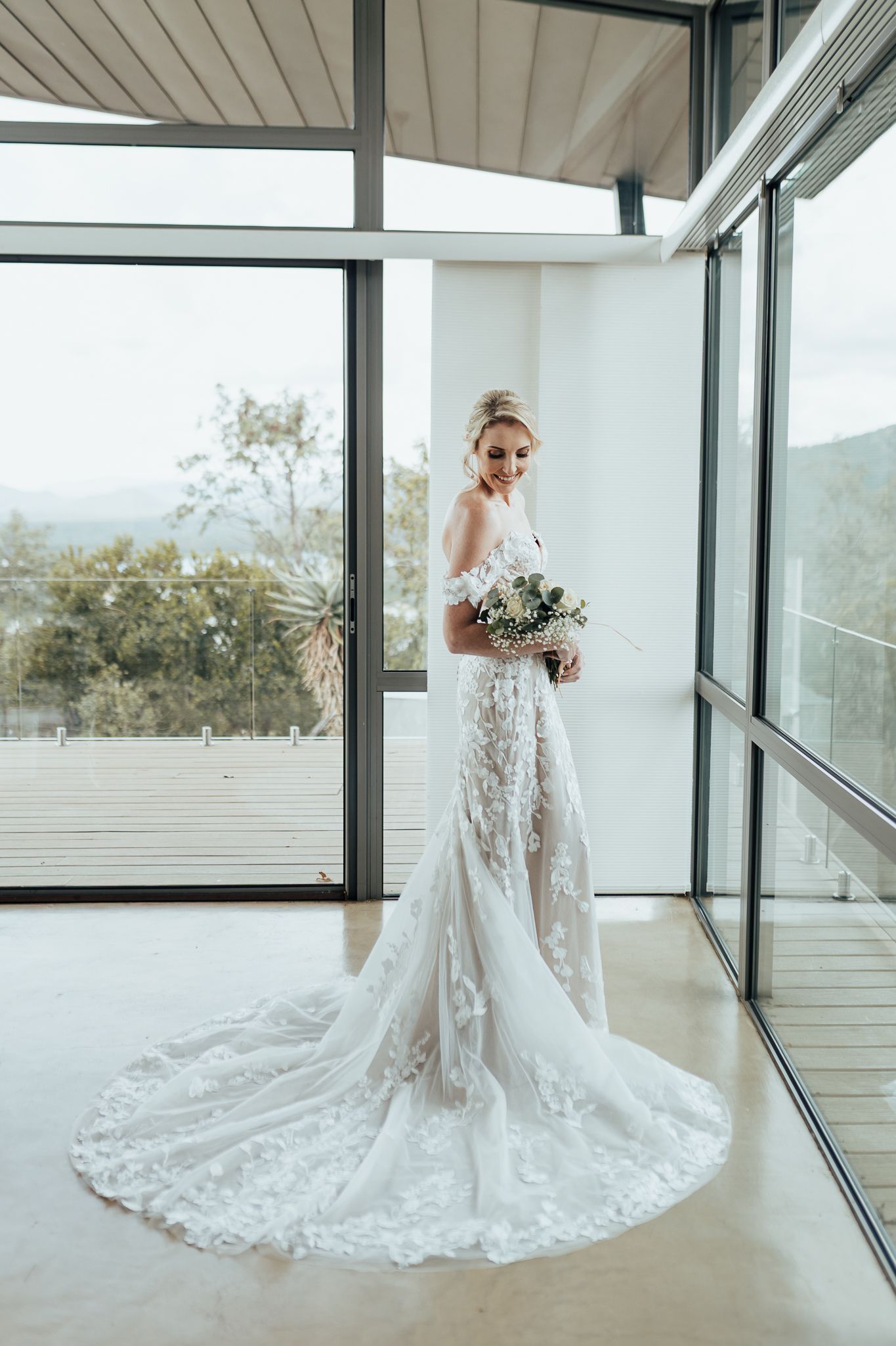 Elegant bride in fitted lace wedding gown with long train poses by floor-to-ceiling windows.