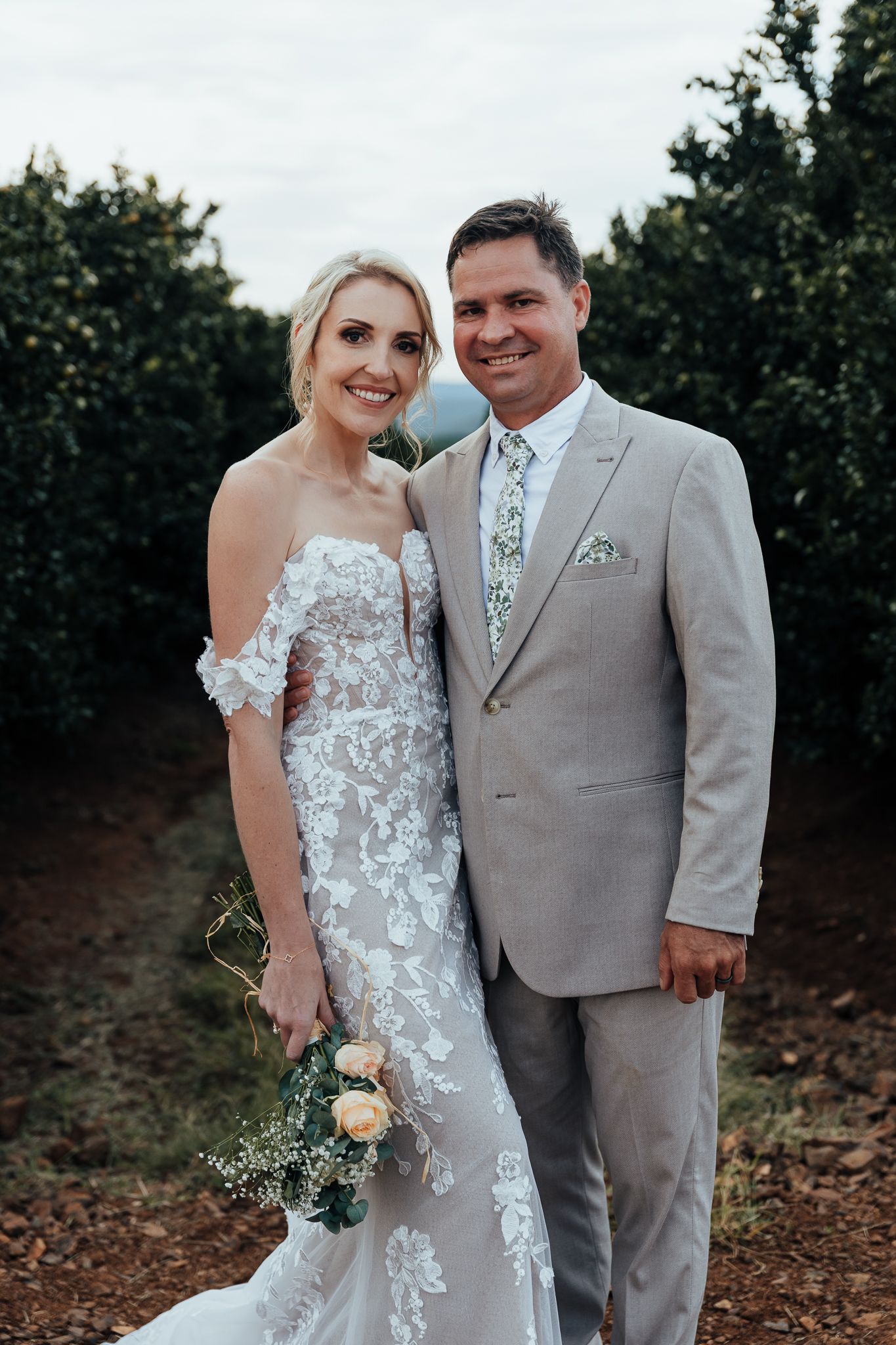 Couple poses together in formal wedding attire against a backdrop of trees.