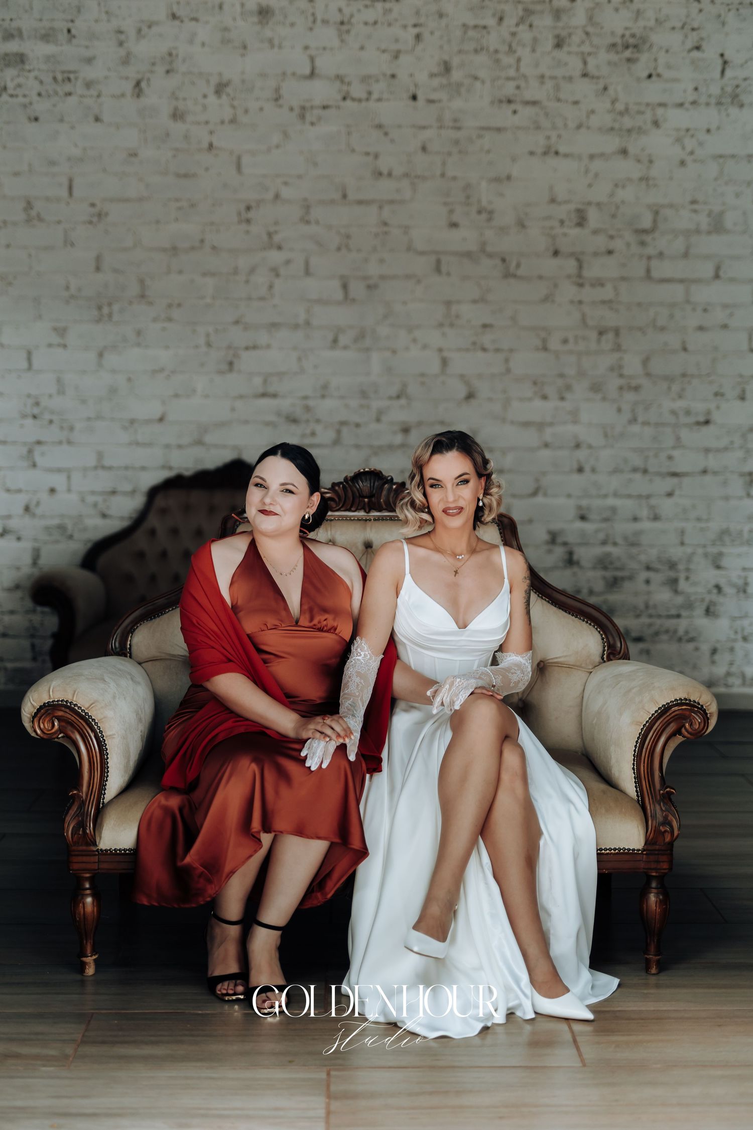Bridesmaids in red dresses sit together on a vintage sofa against white brick wall.