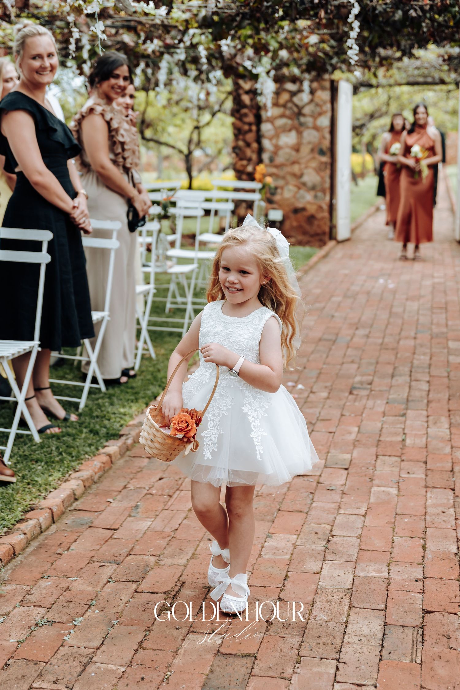 Young flower girl in white dress walking down brick pathway at outdoor wedding ceremony.