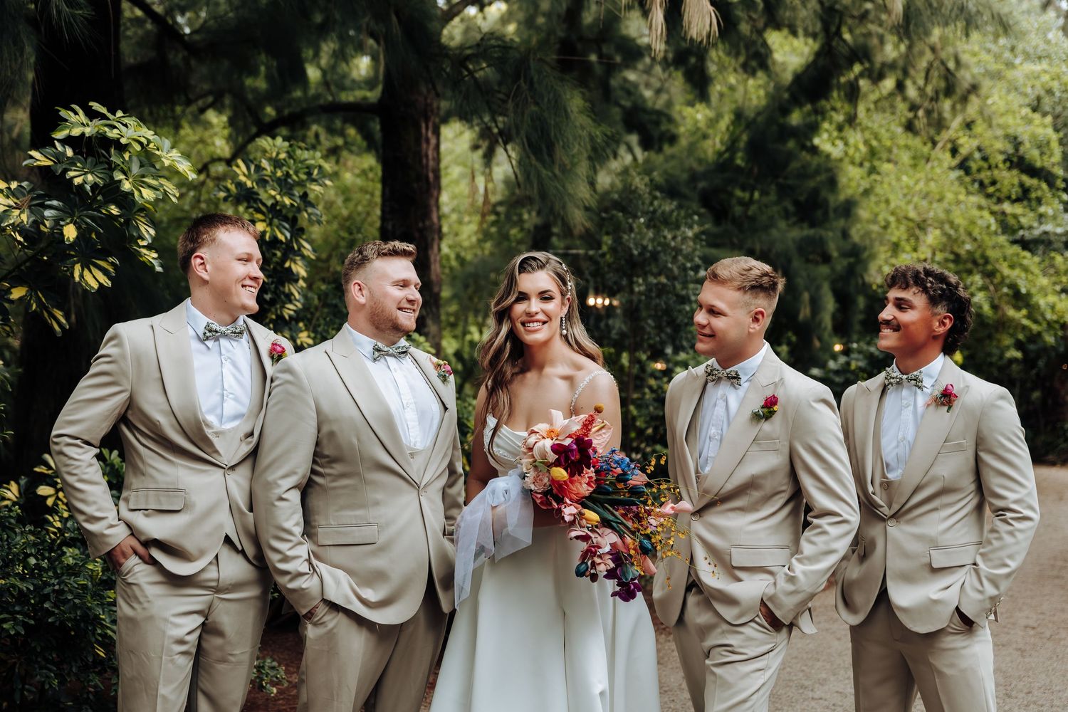 Wedding party members in tan suits and colorful dresses pose together in a lush outdoor garden setting.