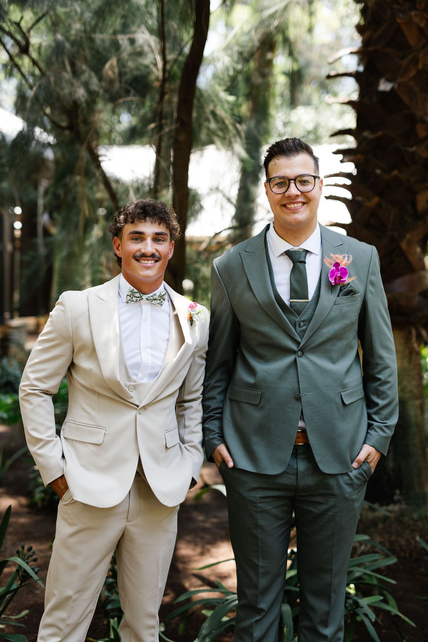 Two people in formal attire, one in beige and one in dark suits, pose together in a garden setting.