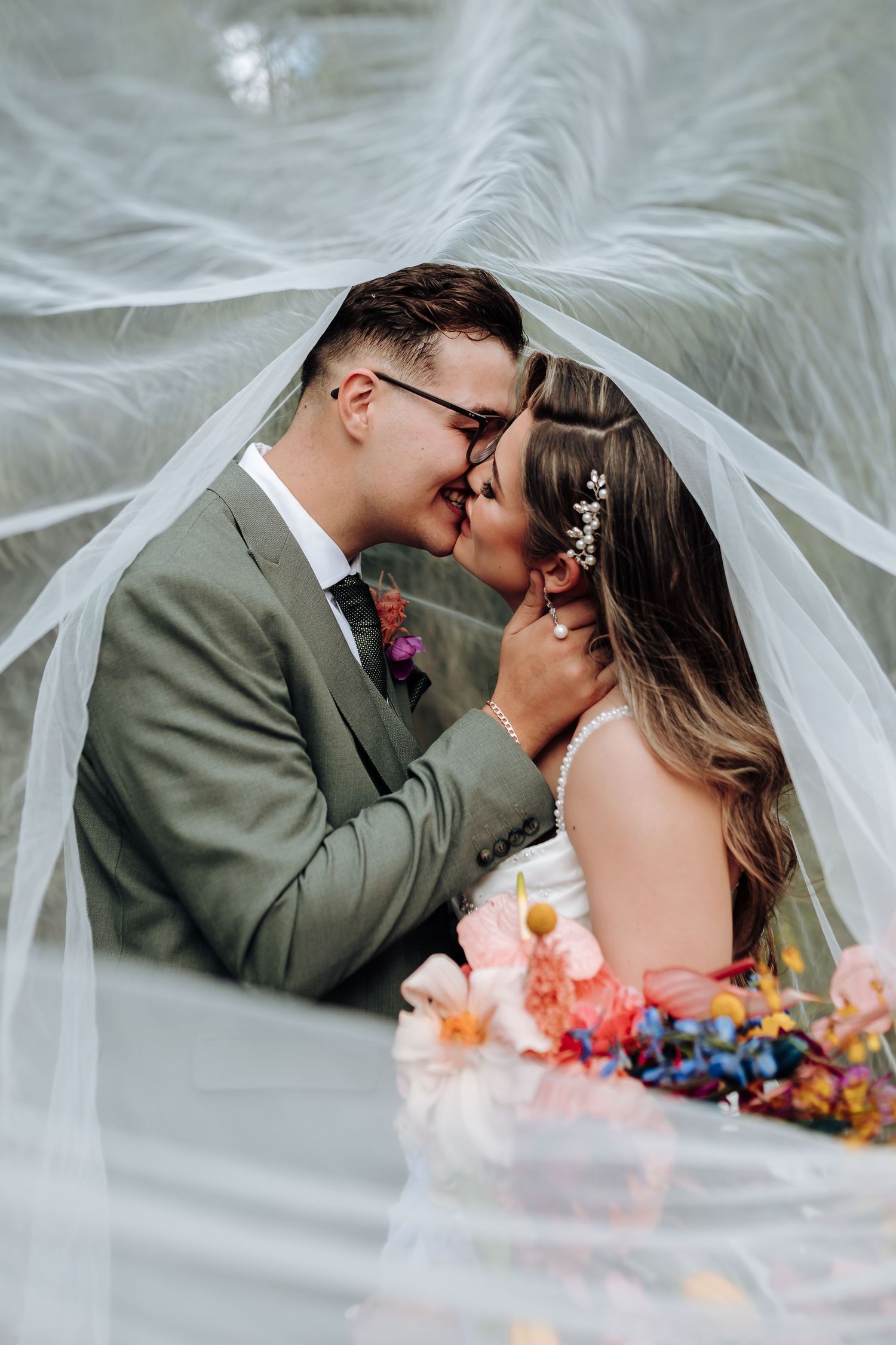 A tender wedding moment captured under flowing white veil with colorful florals in the foreground.