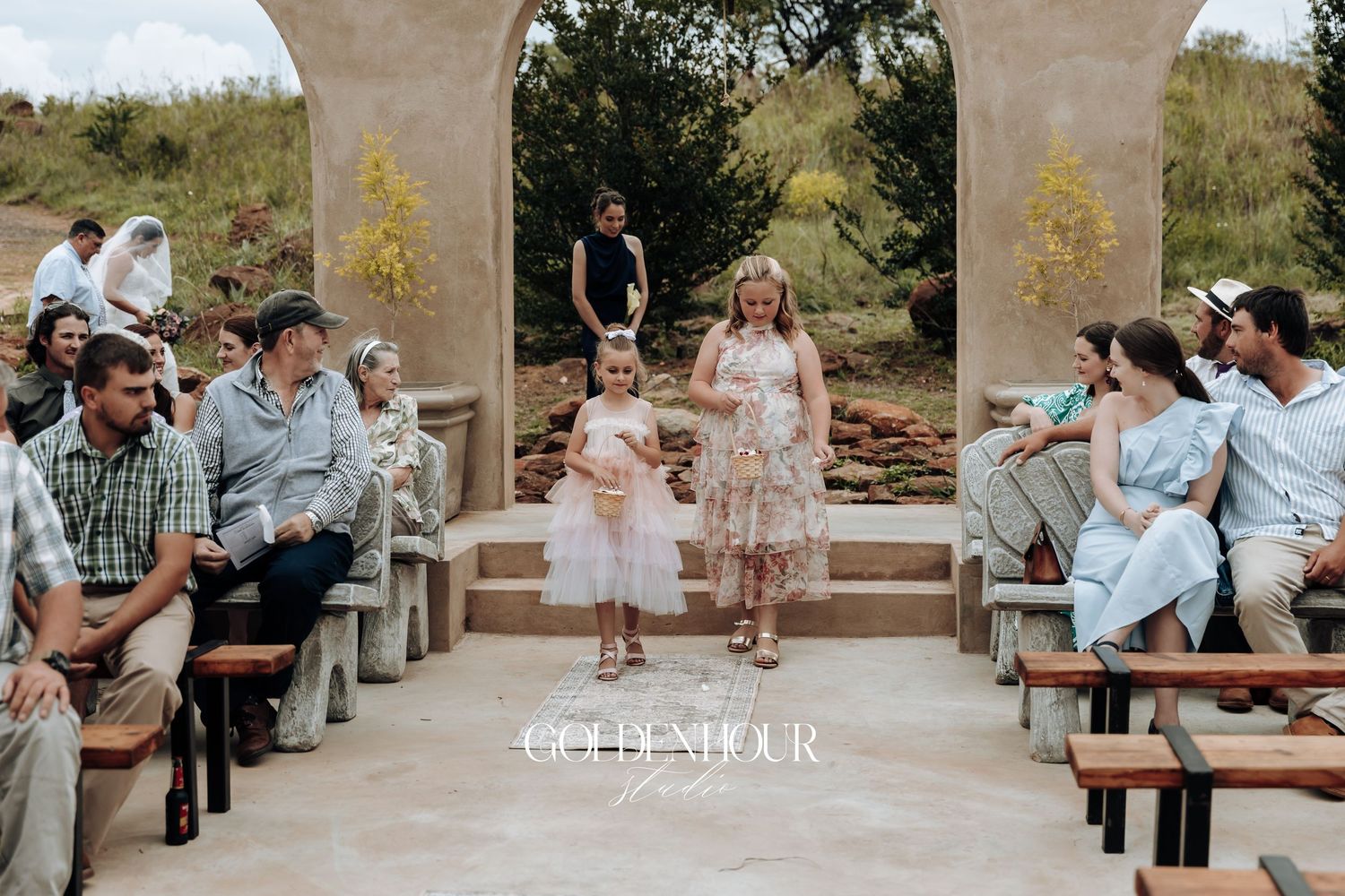 Wedding guests gather on stone benches under arched structures during an outdoor ceremony.