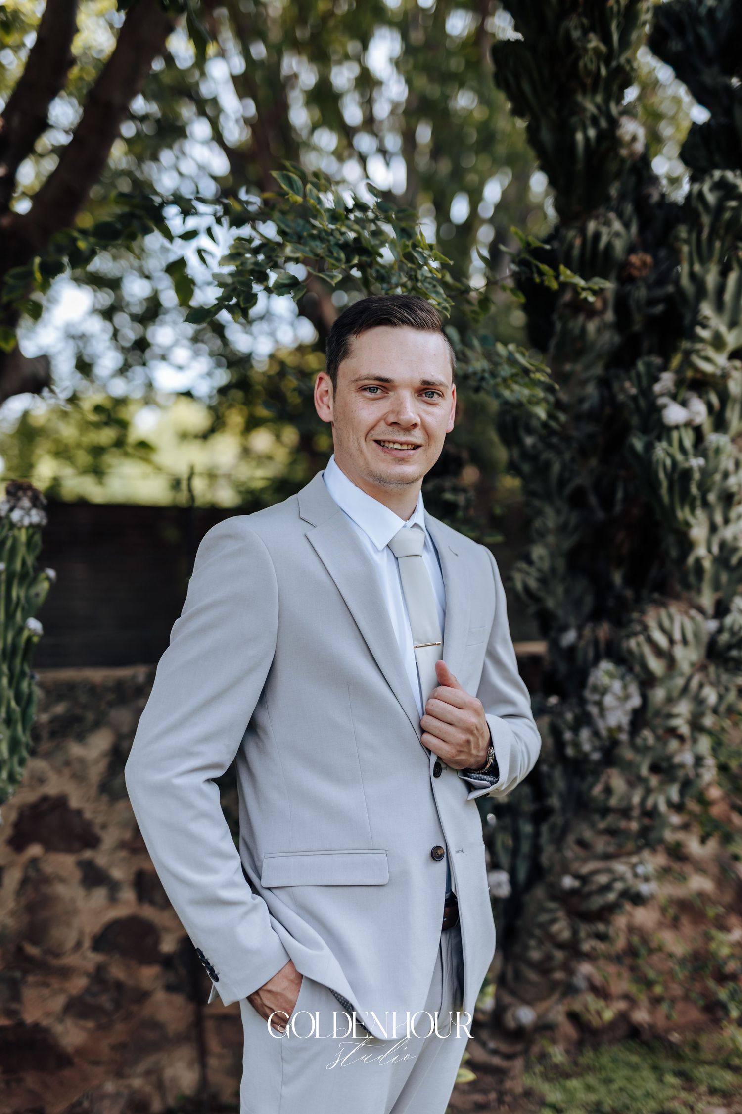 A groom in a light gray suit poses outdoors against a natural backdrop with trees and greenery.