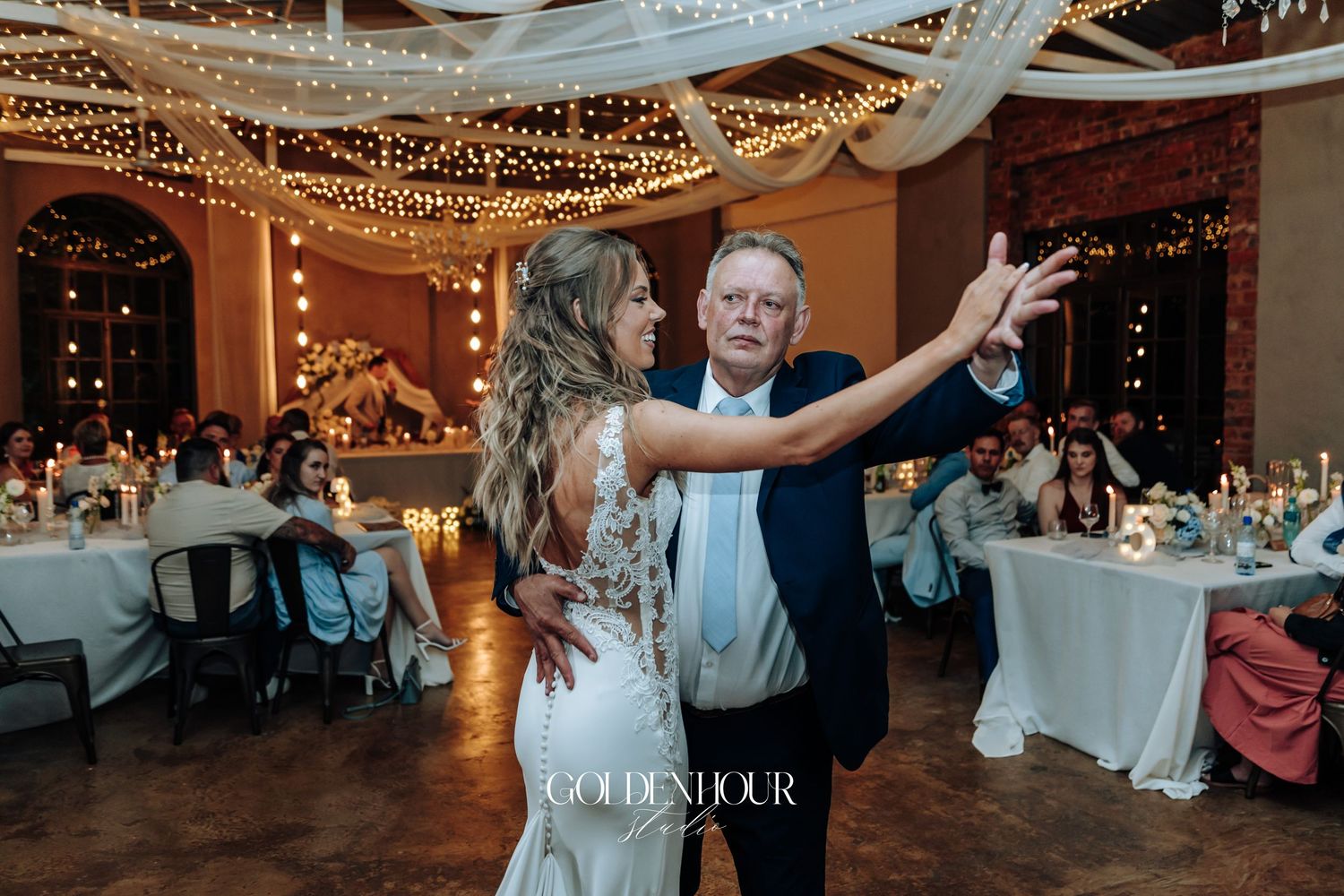 A bride dances with a guest under twinkling lights and white fabric draping at wedding reception.