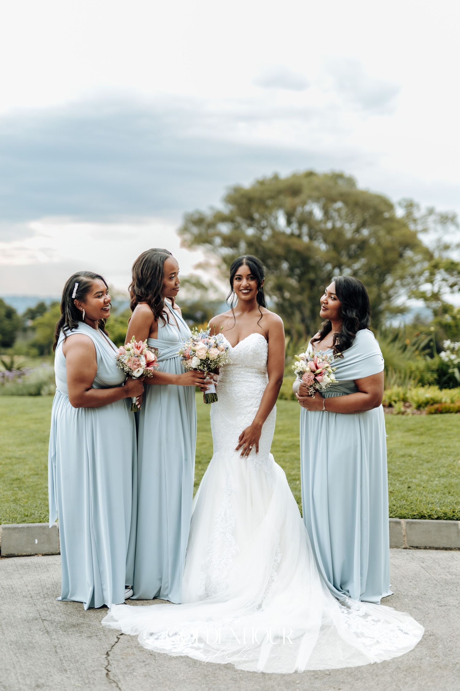 Wedding party poses outdoors wearing elegant light blue bridesmaid dresses with white floral bouquets.