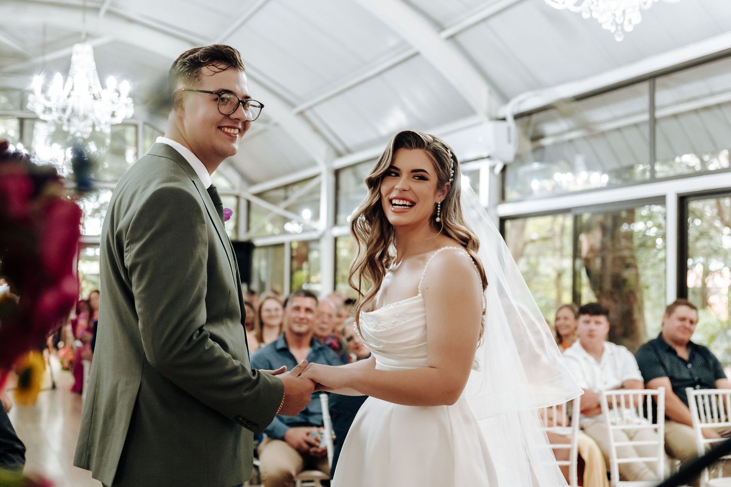 A wedding ceremony taking place in a bright conservatory with elegant chandeliers and natural lighting.