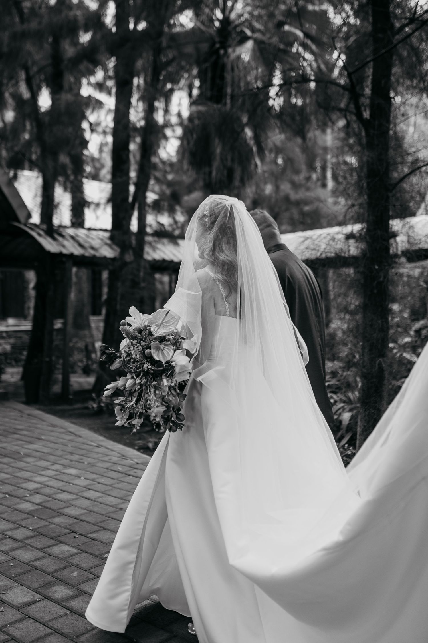 A black and white photo of a bride in a flowing wedding dress holding a bouquet walking along a stone path.