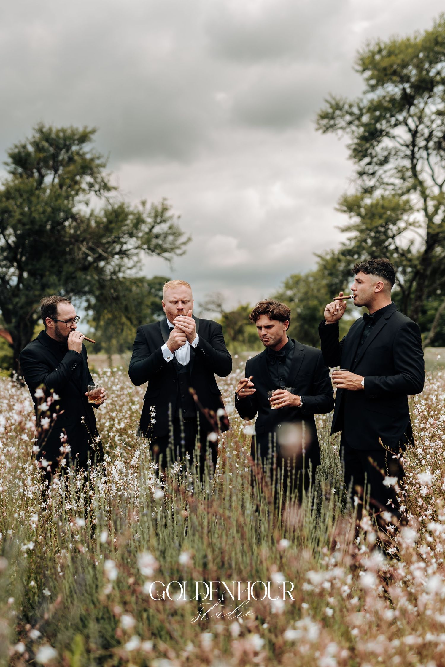 A group in black attire shares drinks together in a field of white wildflowers under cloudy skies.