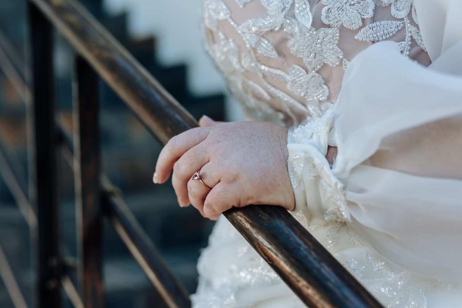 Close-up view of a hand wearing a wedding ring while holding an ornate metal railing, with white lace fabric visible.