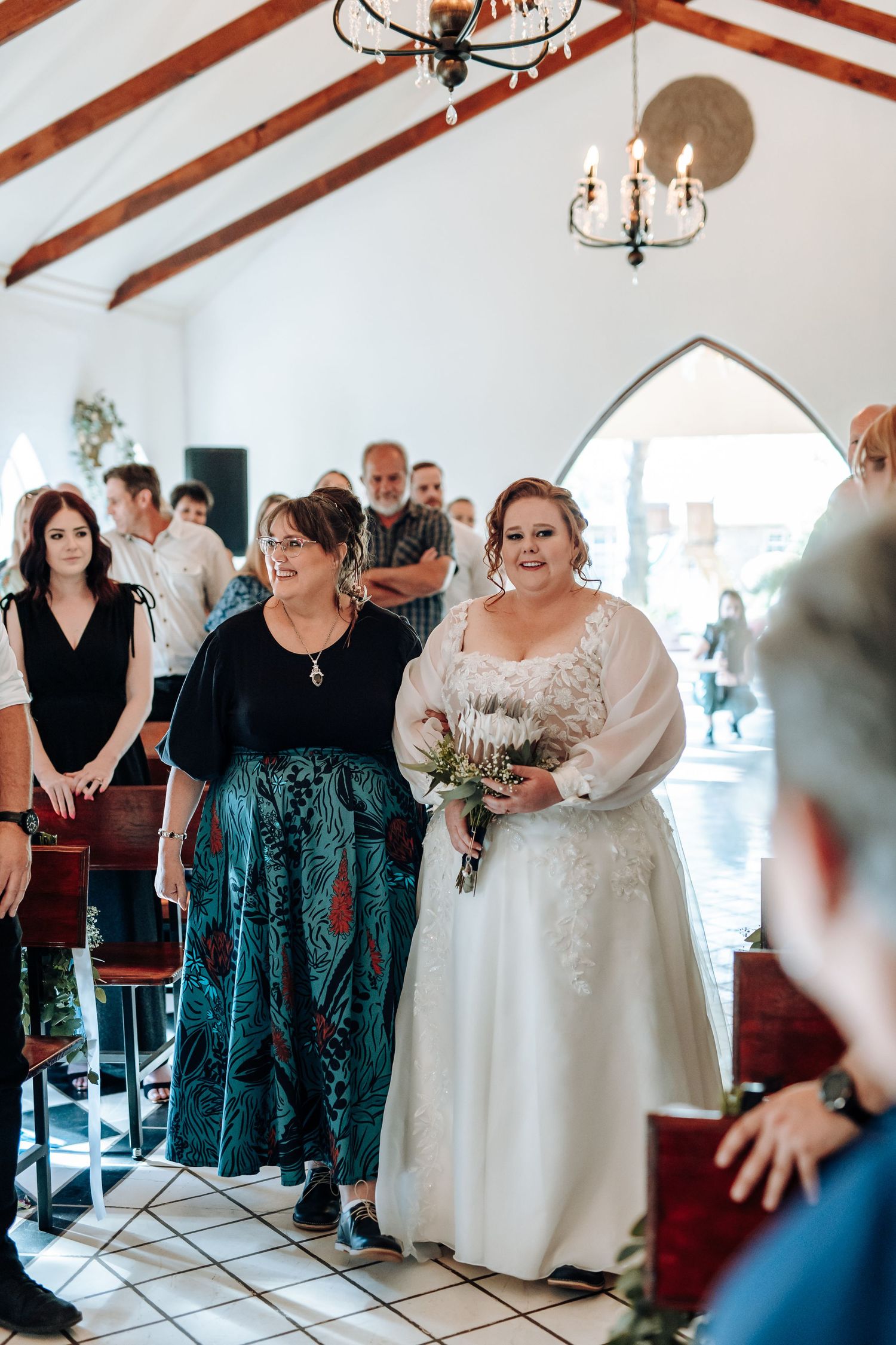 Wedding party members in teal dresses and white gowns gather for photos in an elegant venue with vaulted ceilings.