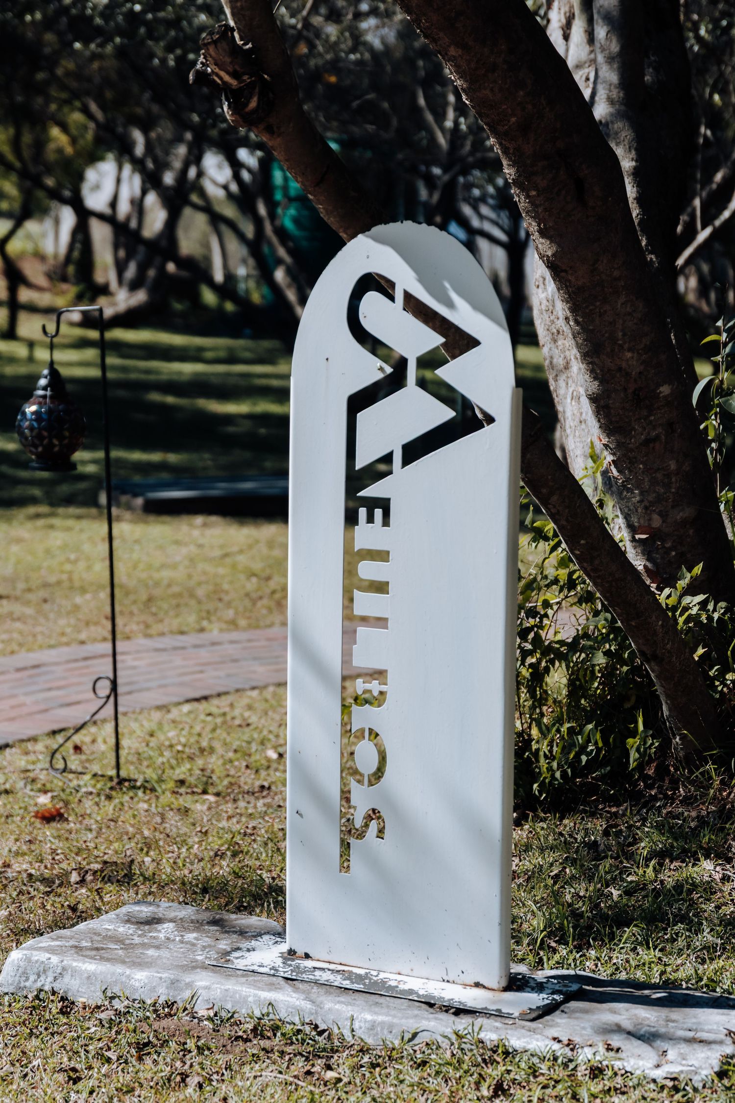 White arched sign for Melrose property standing on concrete base in lush green garden near large tree.