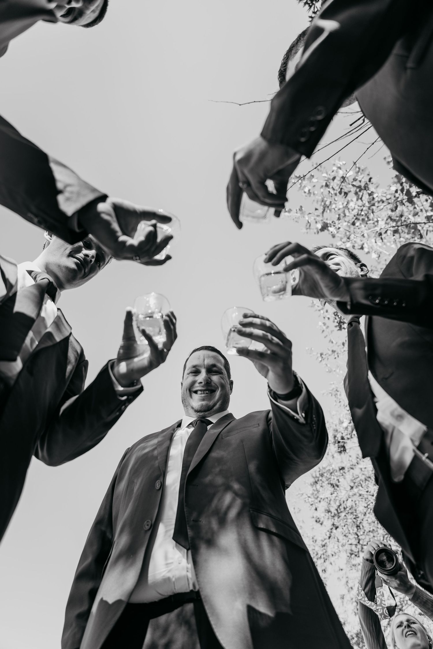 Black and white photo of groom toasting with groomsmen, shot from below with sky background.