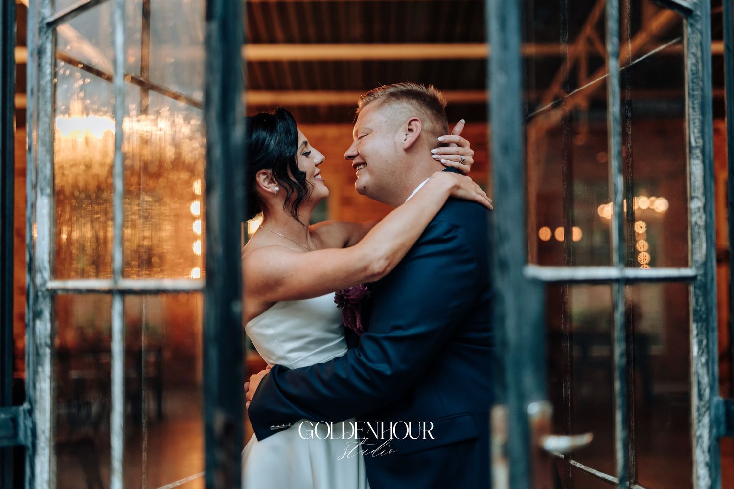 Bride and groom sharing a joyful embrace framed by ornate glass doors with warm lighting.