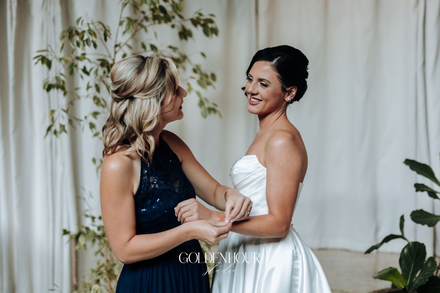 Bridesmaid in navy sequin dress helping bride fasten her white wedding gown before the ceremony.
