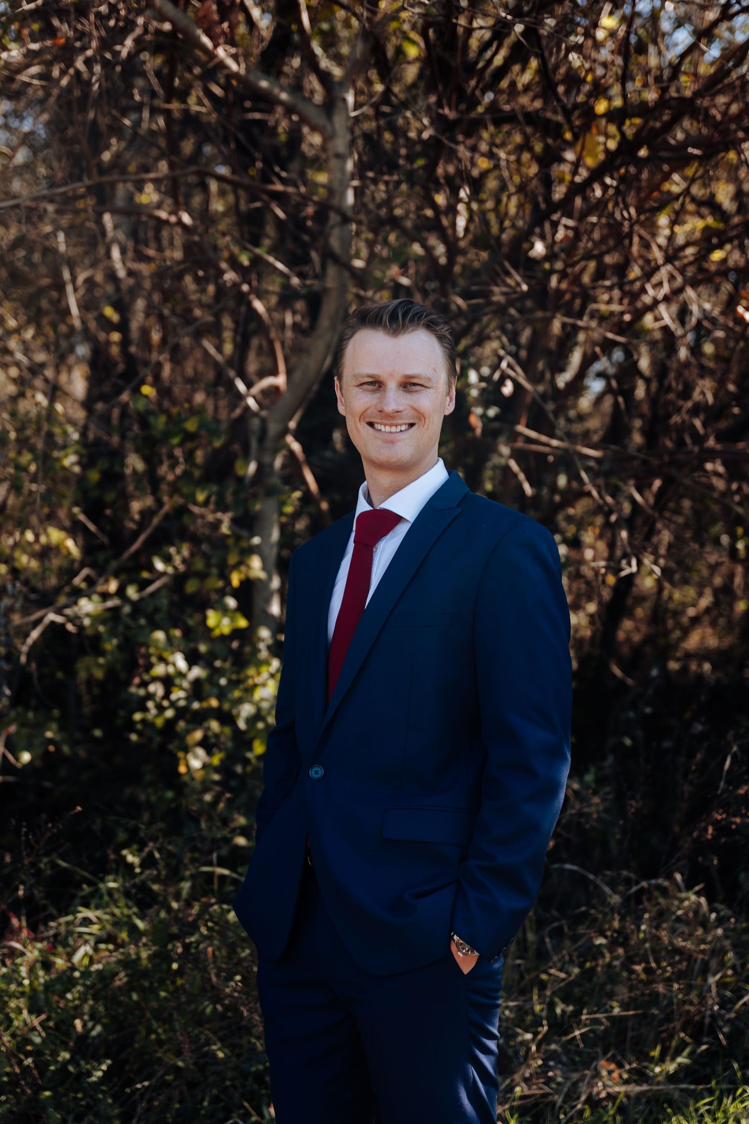 Smiling man in navy blue suit and red tie standing outdoors among autumn trees.