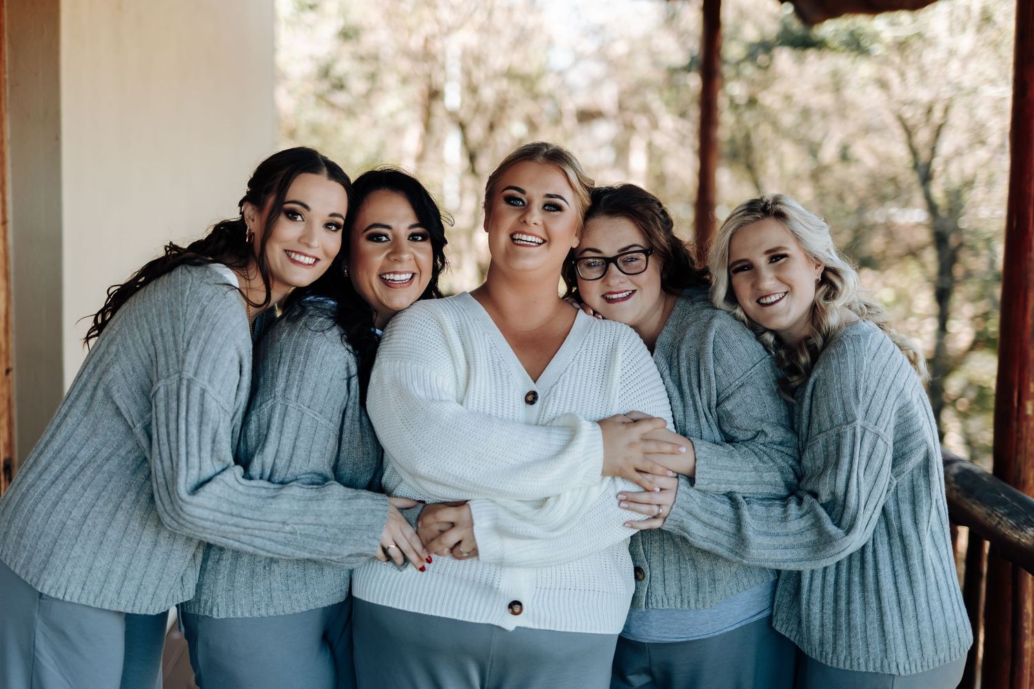 Five women in matching gray sweaters and pants smile together, with the bride in a white cardigan centered among her bridesmaids.