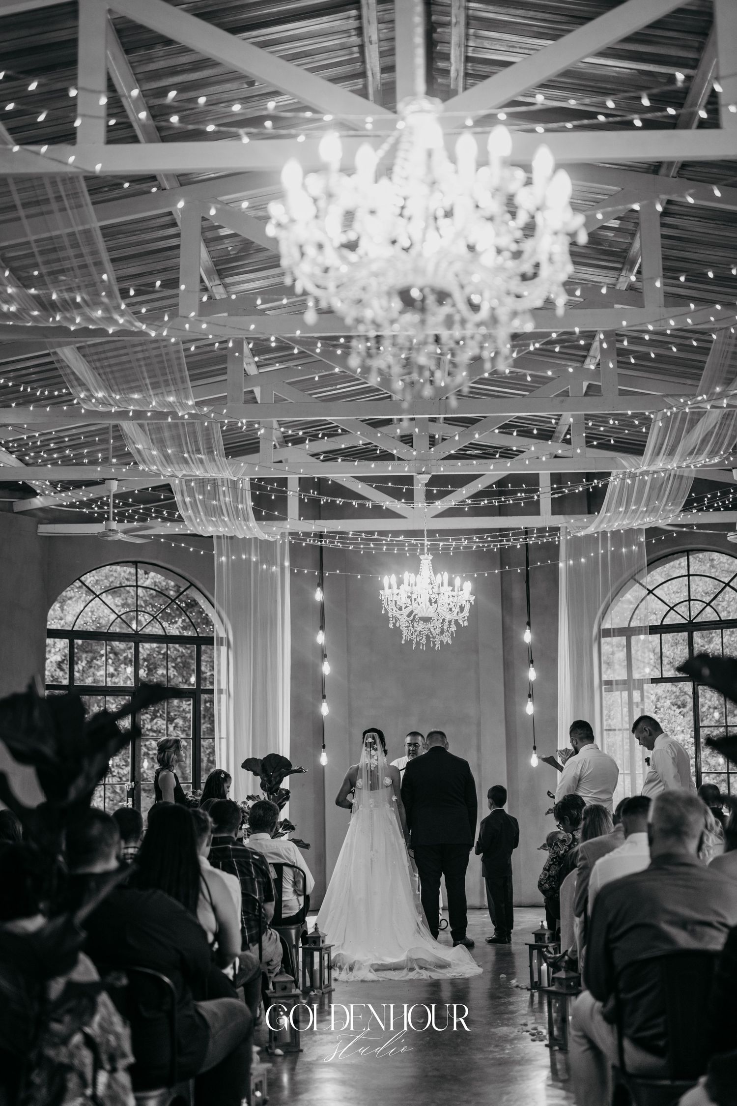 Black and white wedding ceremony under crystal chandeliers and fairy lights in an elegant glass-roofed venue.