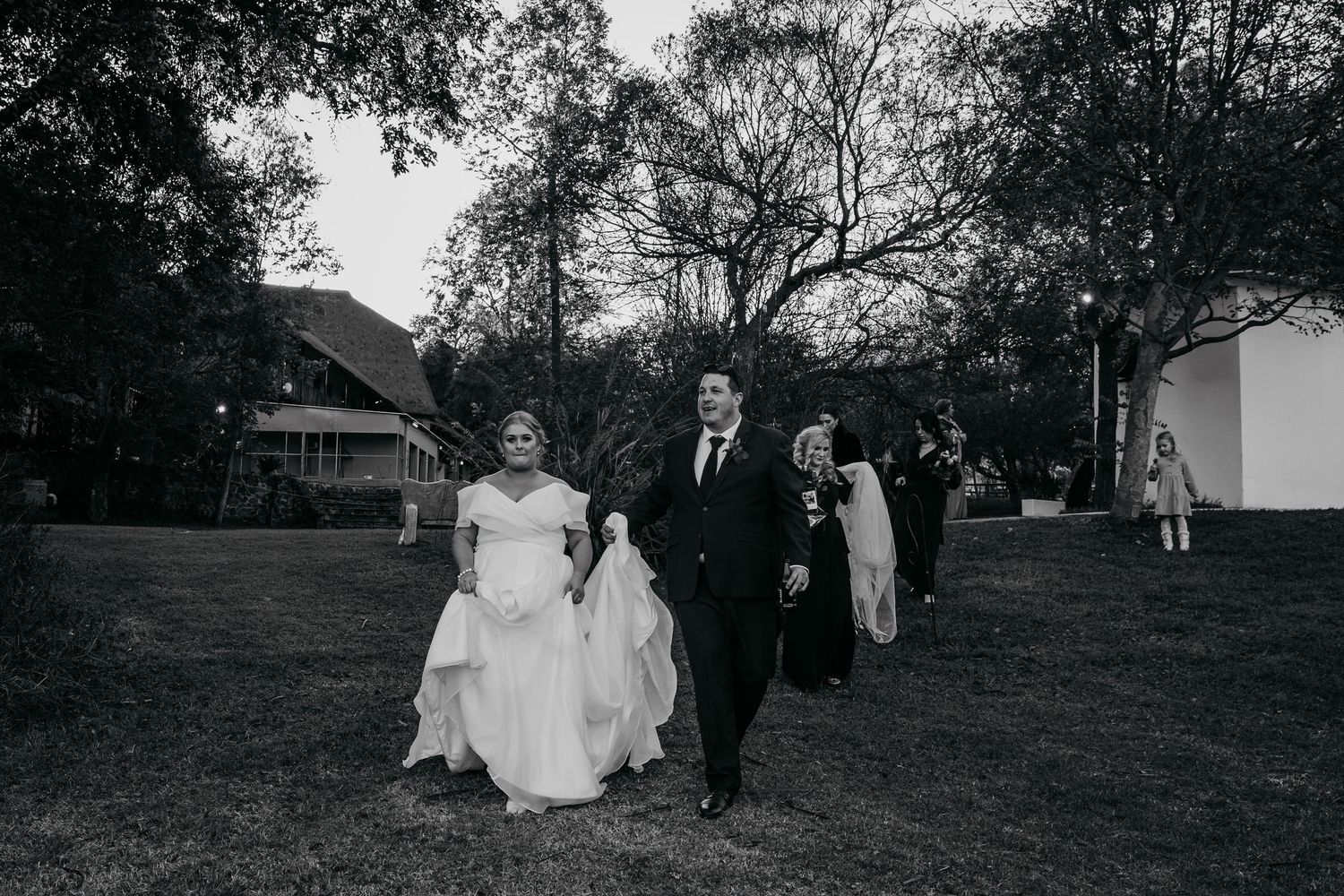 Black and white photo of a bride and groom walking across a lawn with wedding guests following behind them.
