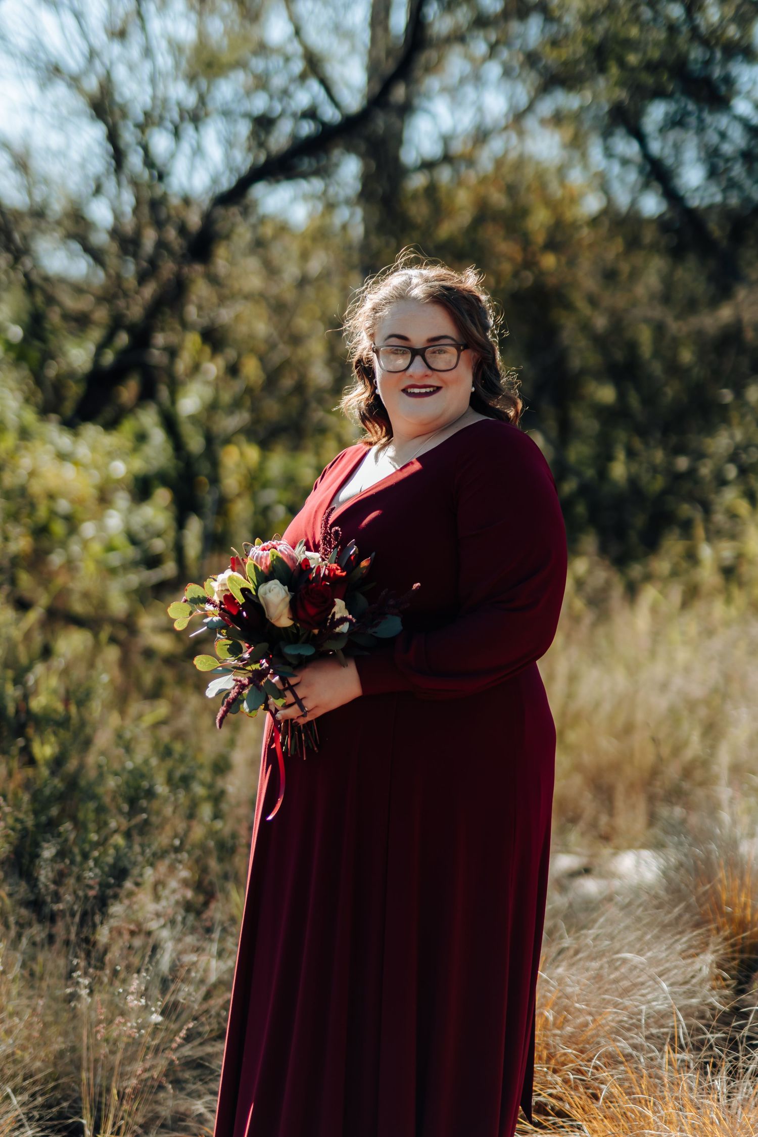 Woman in burgundy gown holding floral bouquet, smiling outdoors in natural bush setting.