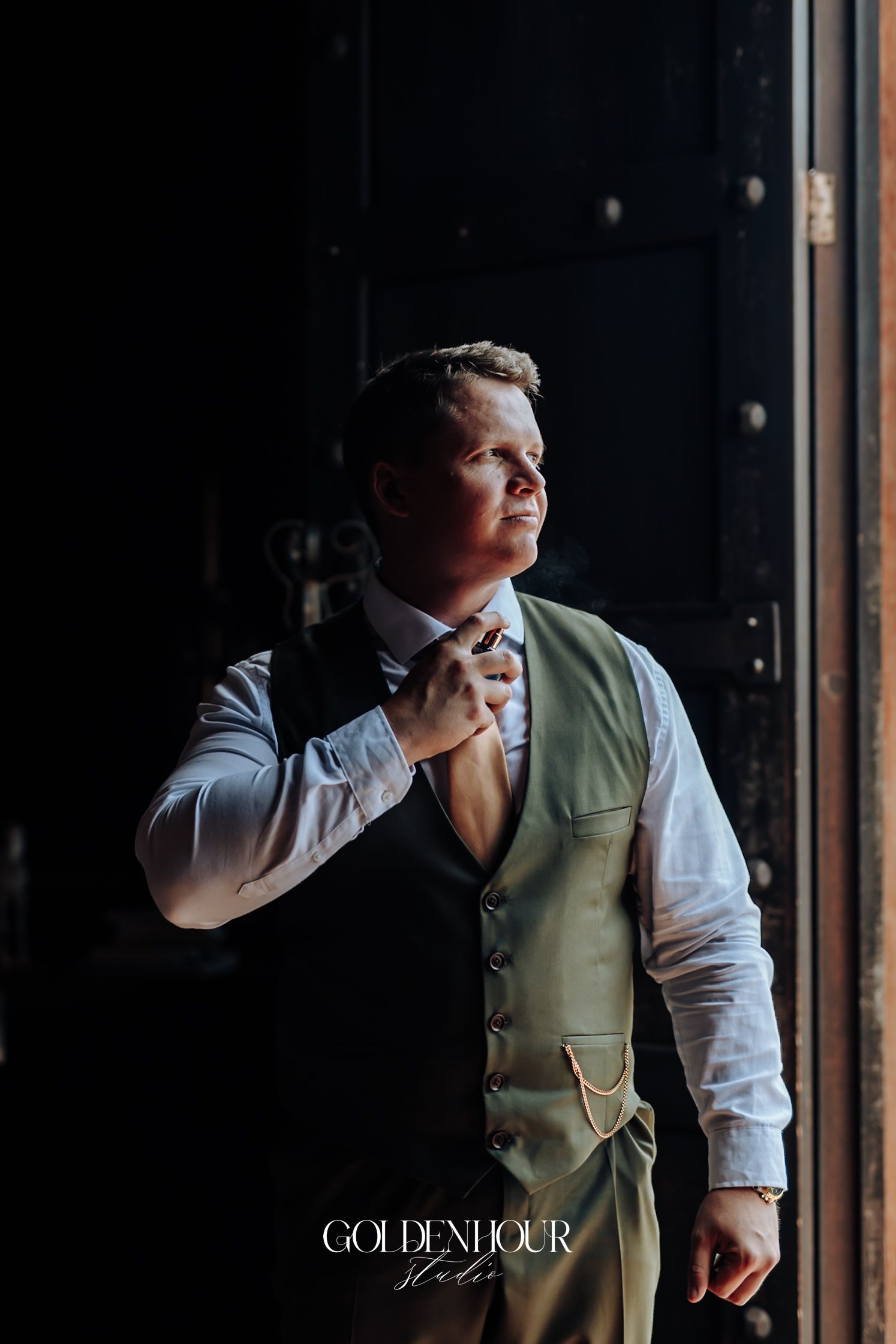 Groom in sage green vest adjusting tie near dramatic dark doorway with golden light.