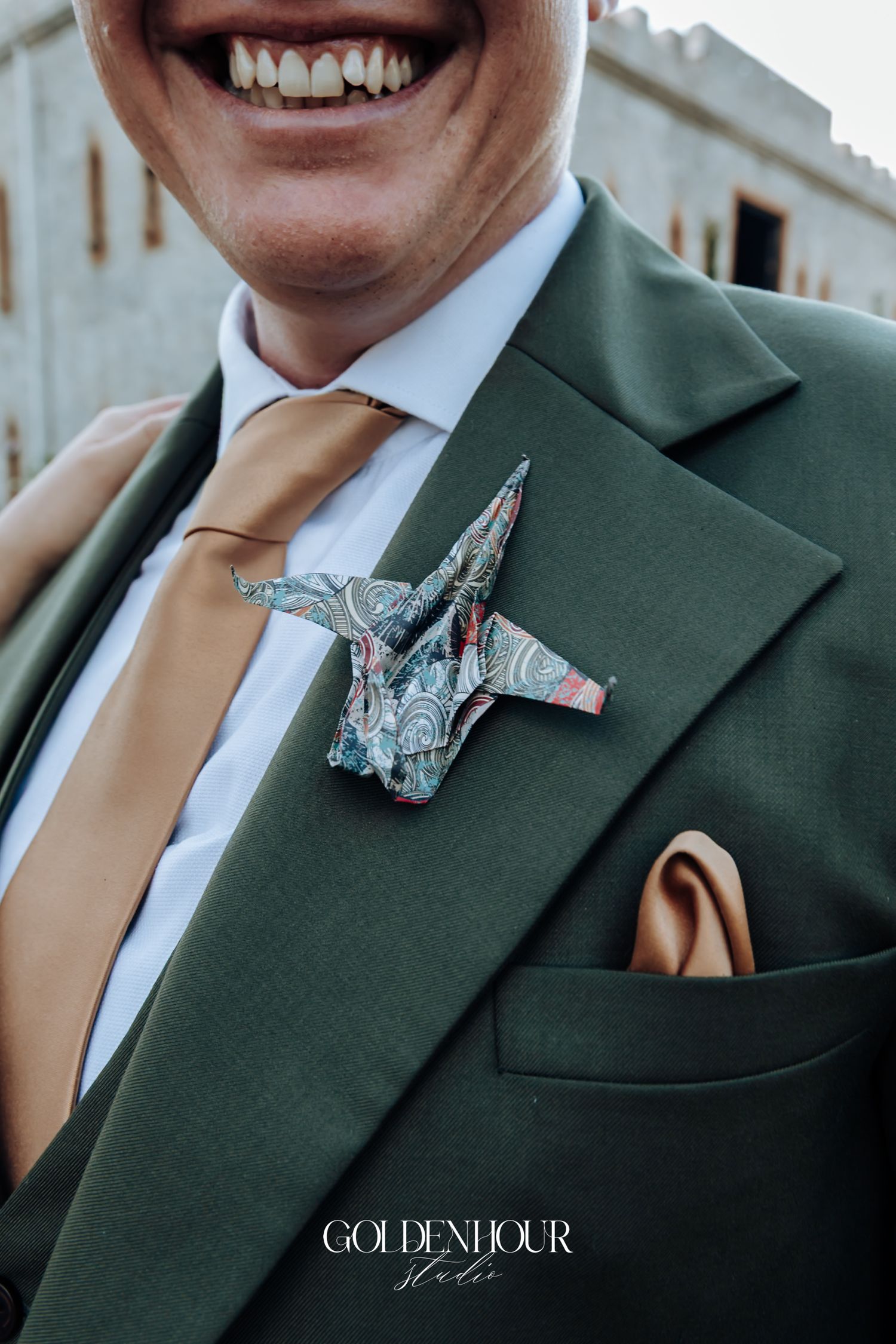 Groom in green suit with gold tie, origami crane boutonniere, and gold pocket square, smiling at wedding.