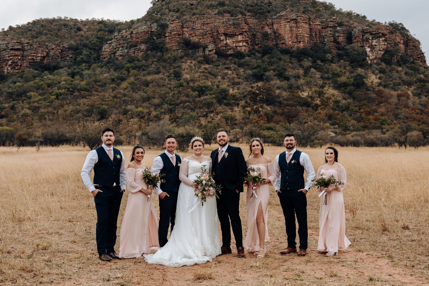 A wedding party stands together in a grassy field with a dramatic red rock mountain cliff backdrop.