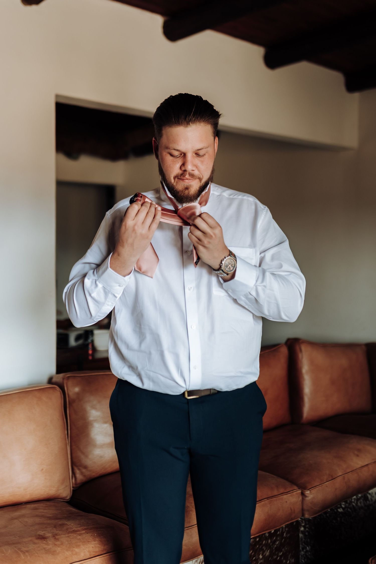 Man in white dress shirt adjusts his necktie while standing in front of brown leather couch.